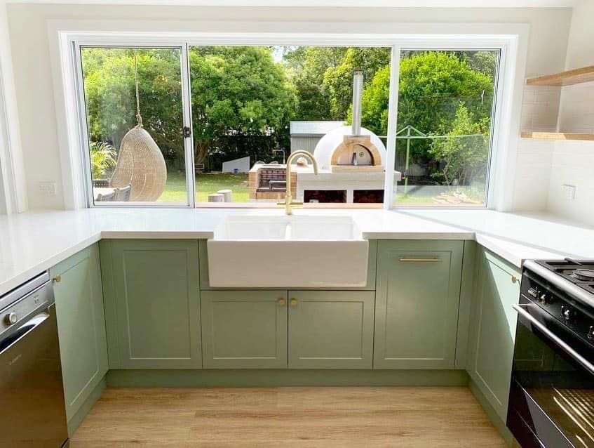 A kitchen with green cabinets , a white sink , a stove , and a large window.