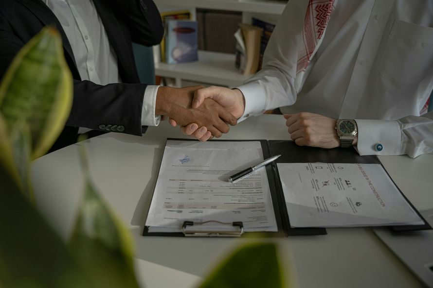 Two people shaking hands over signed documents on a desk in an office