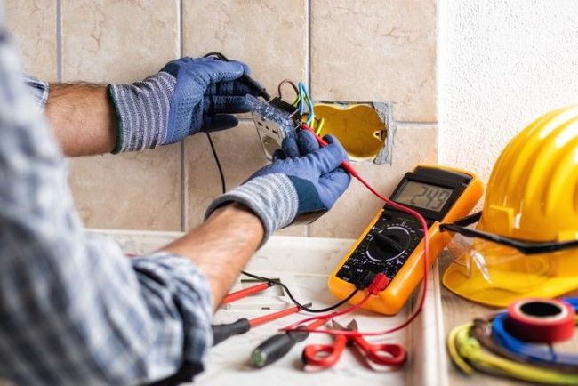 Electrician in blue gloves tests wires in a wall outlet, using a multimeter; yellow hard hat nearby.