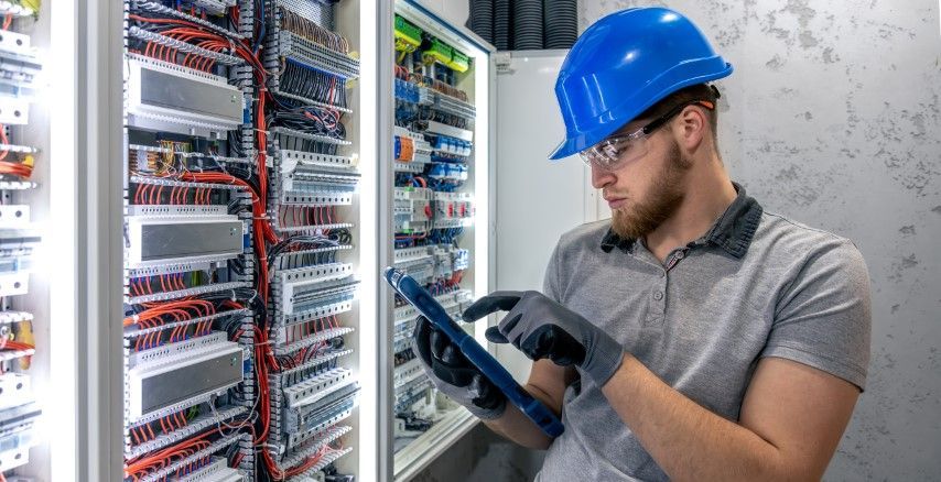 Electrician wearing a blue hard hat and gloves inspects wiring in a panel, using a tablet.