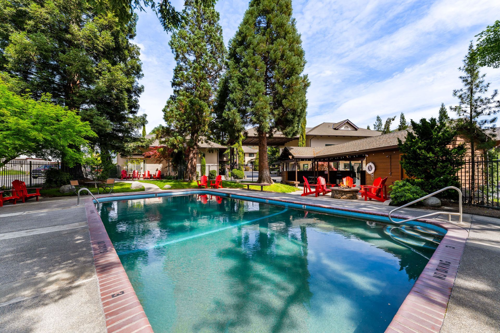Outdoor swimming pool surrounded by tall trees, lounge chairs, and a brick patio at a resort or apartment complex