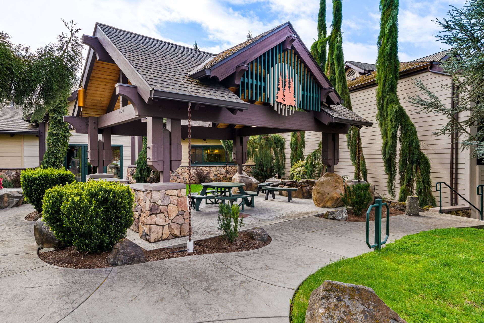 Covered picnic shelter with wooden beams, stone columns, benches, and landscaped greenery in a park-like setting