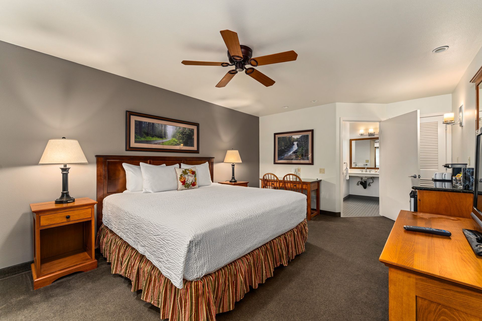 Hotel bedroom with king bed, ceiling fan, lamps, dresser, desk, and open bathroom doorway