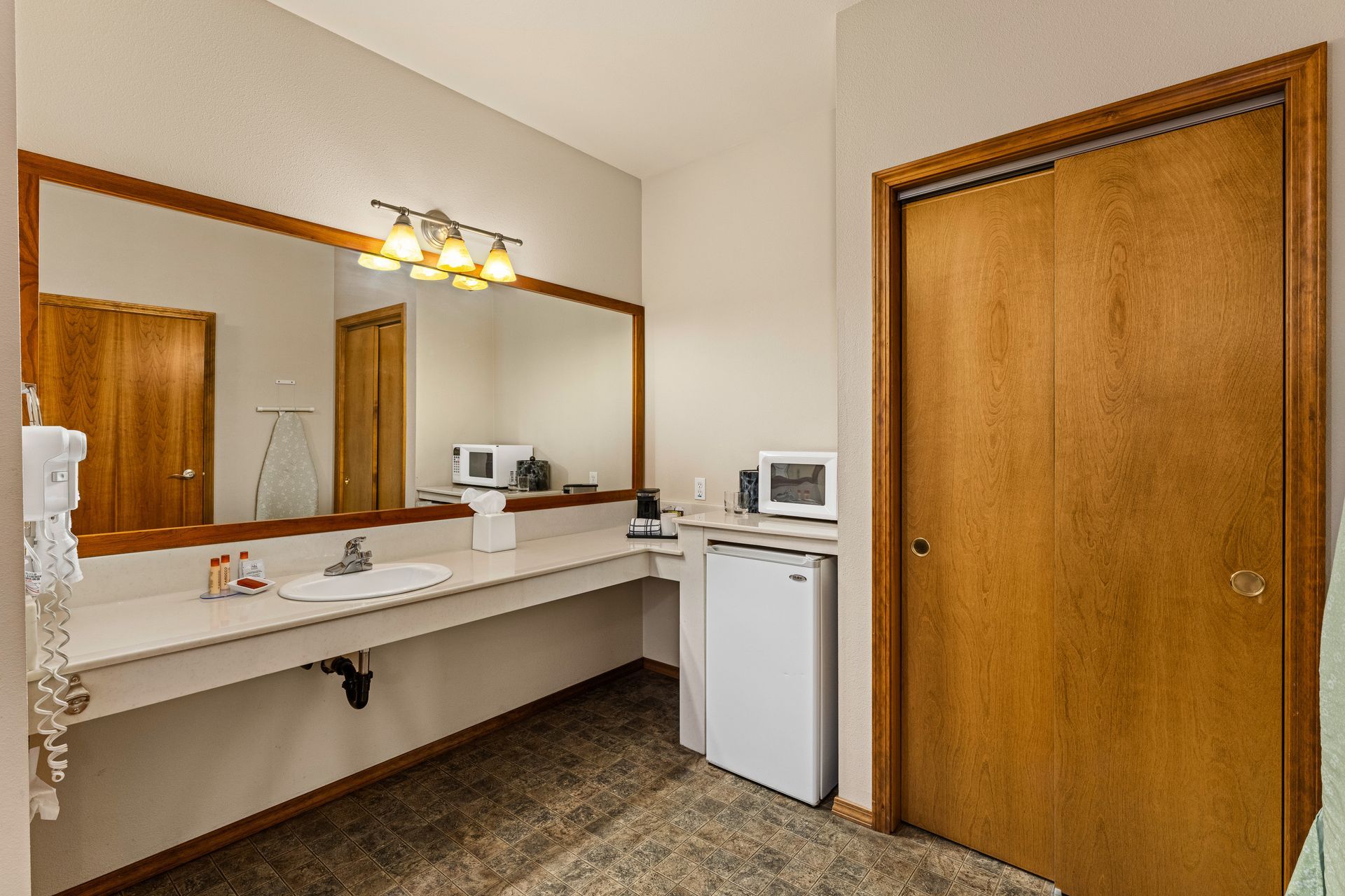 Small motel-style bathroom vanity with mirror, sink, mini fridge, microwave, and wooden door