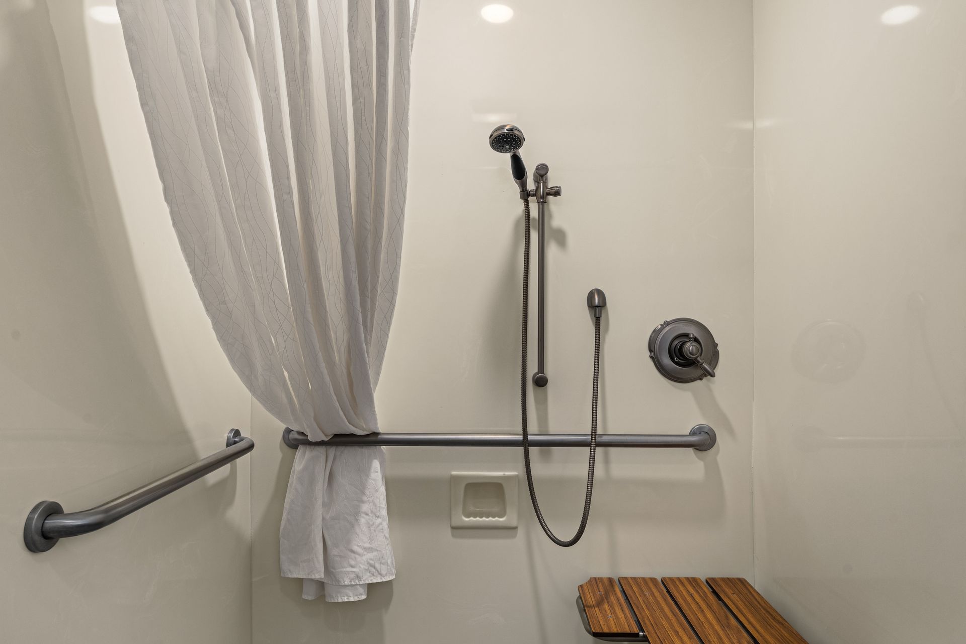 Accessible shower with grab bars, handheld showerhead, curtain, and wooden bench.
