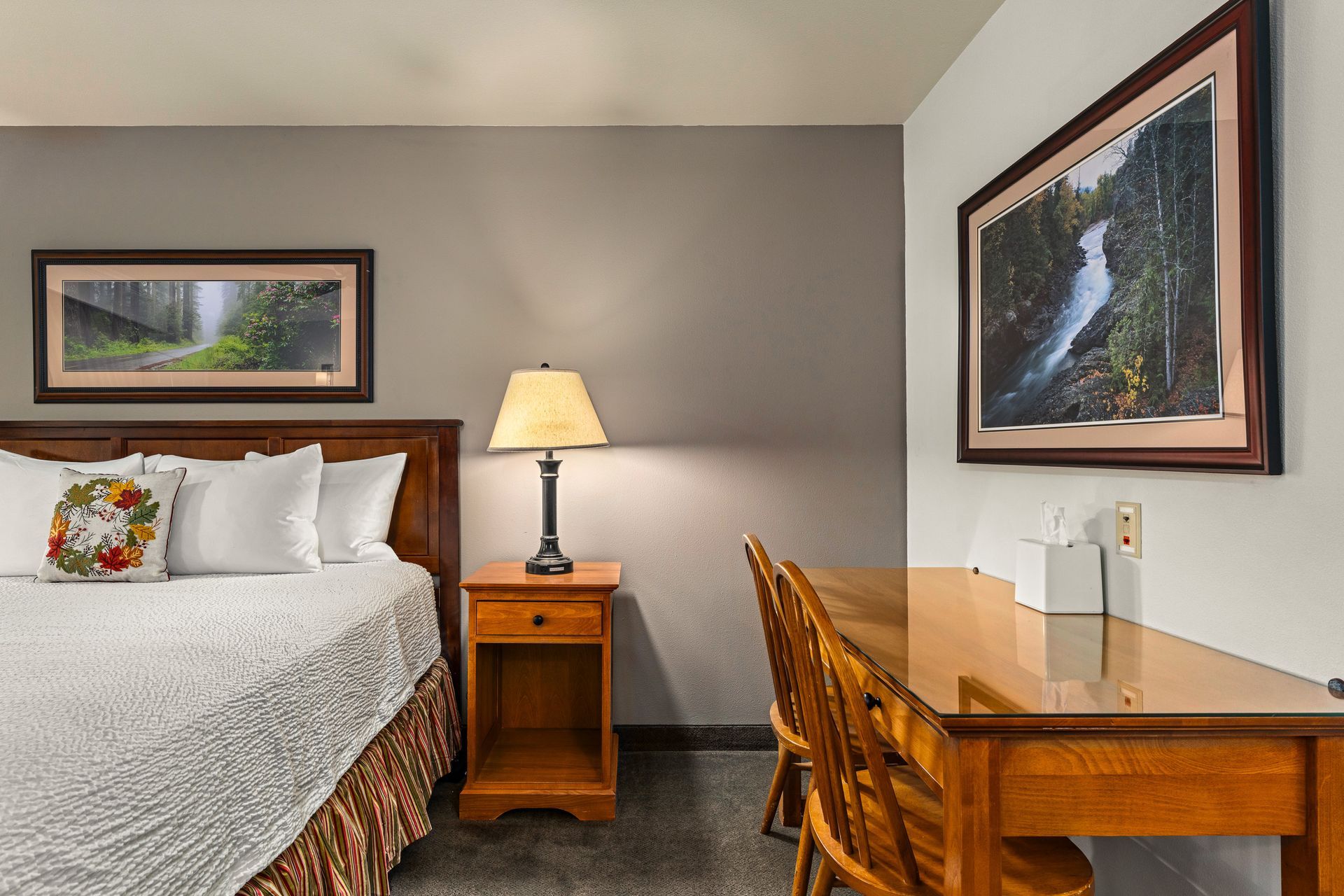 Small hotel room with twin bed, bedside lamp, wooden desk, and framed wall art.