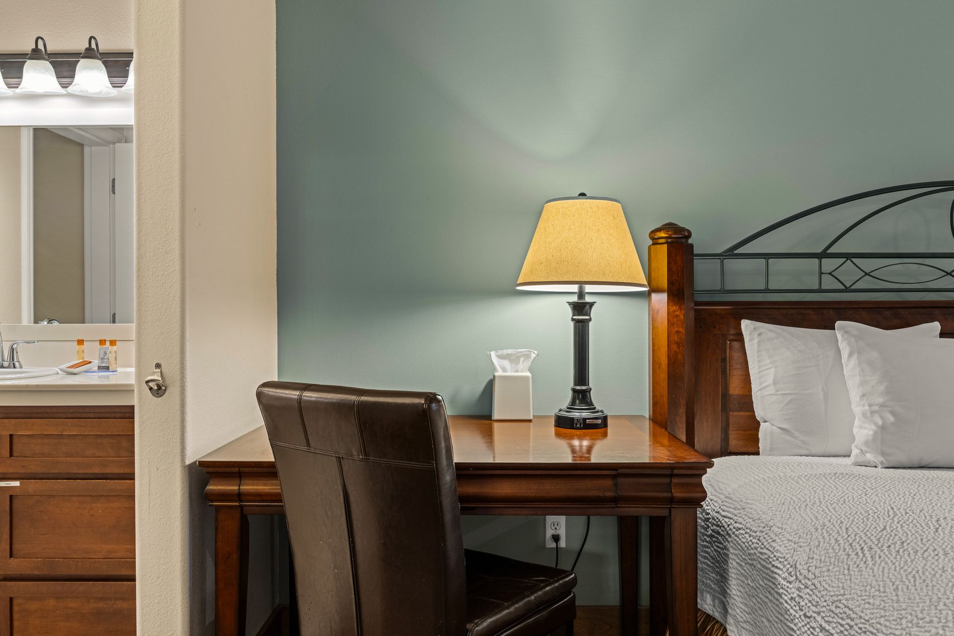 Hotel room desk with lit lamp beside a bed and bathroom vanity nearby