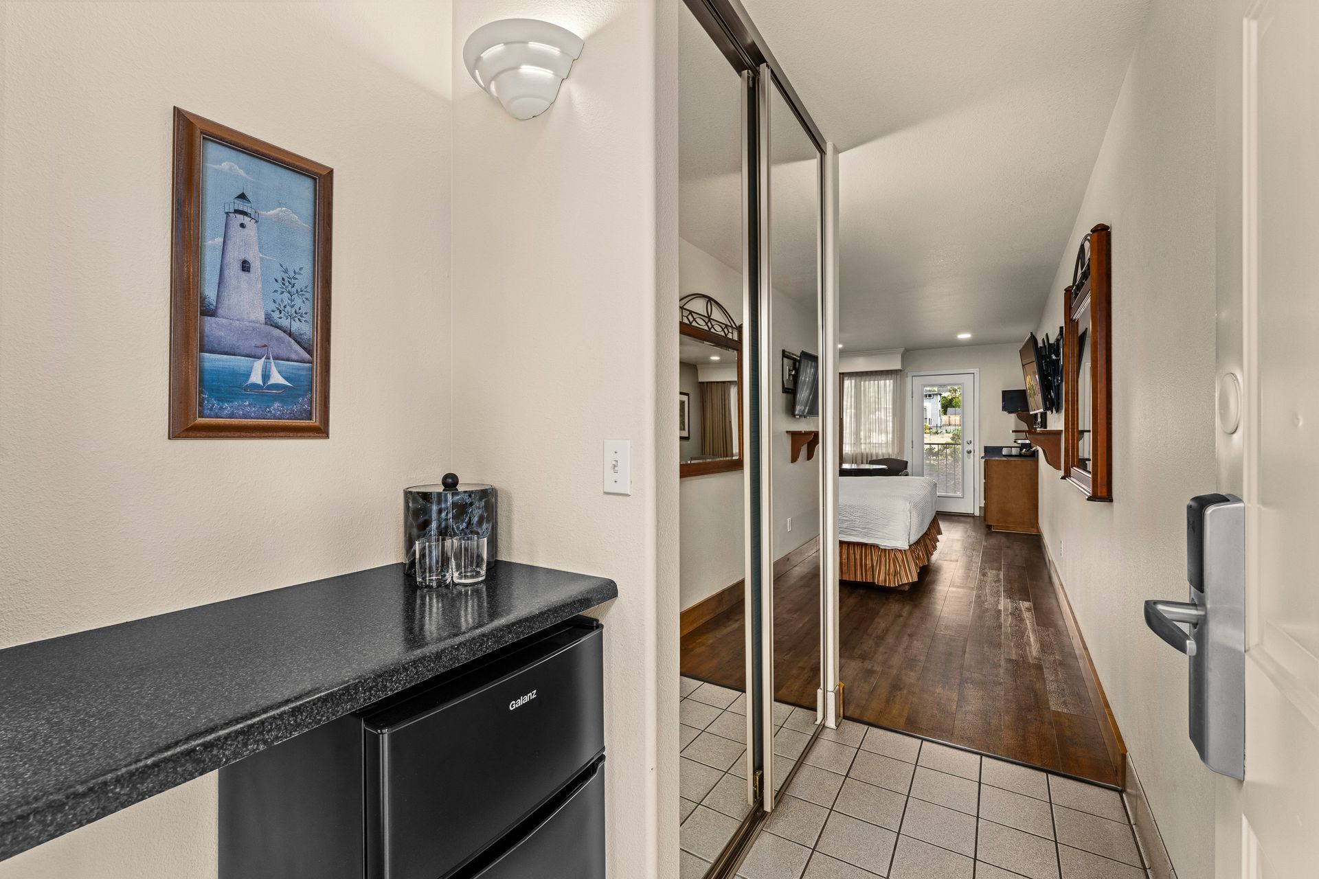 Narrow apartment hallway with tiled floor, black countertop, and a view into a bedroom and living area.