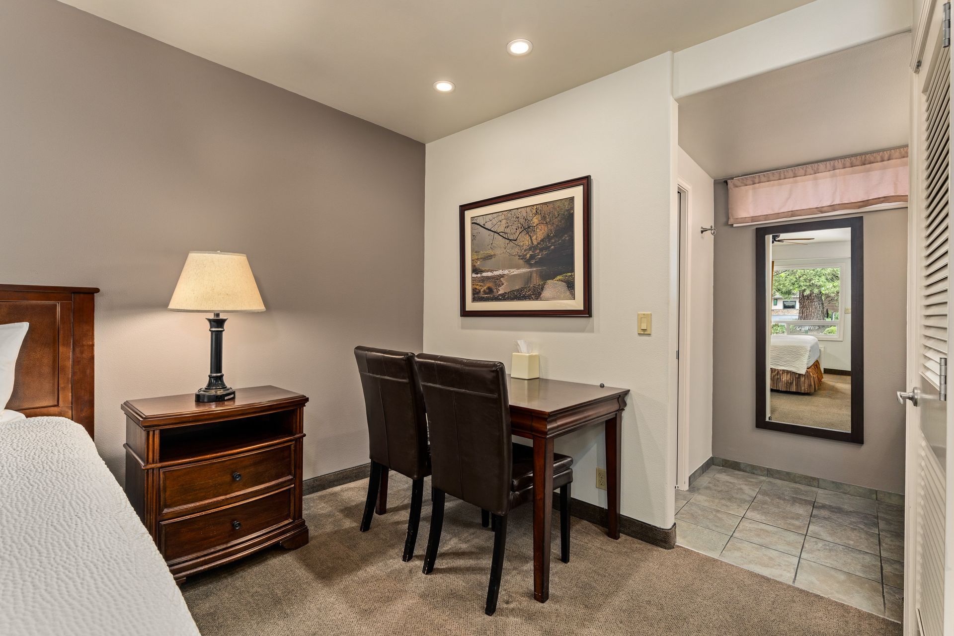 Small bedroom nook with desk and chairs, lamp on nightstand, carpet, and hallway with tiled floor.