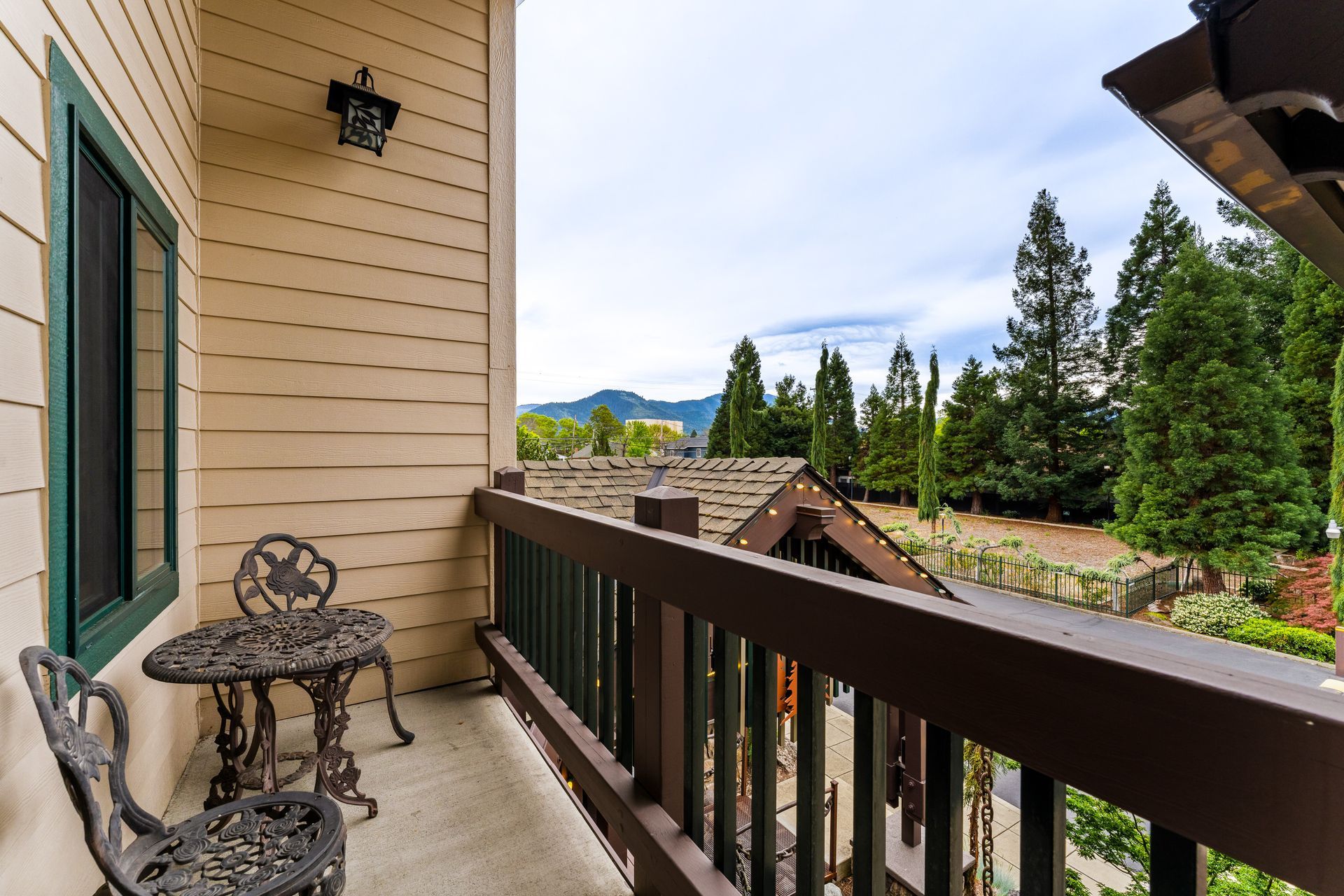 Balcony with wrought-iron chairs, tan siding, and a view of trees, rooftops, and distant mountains.