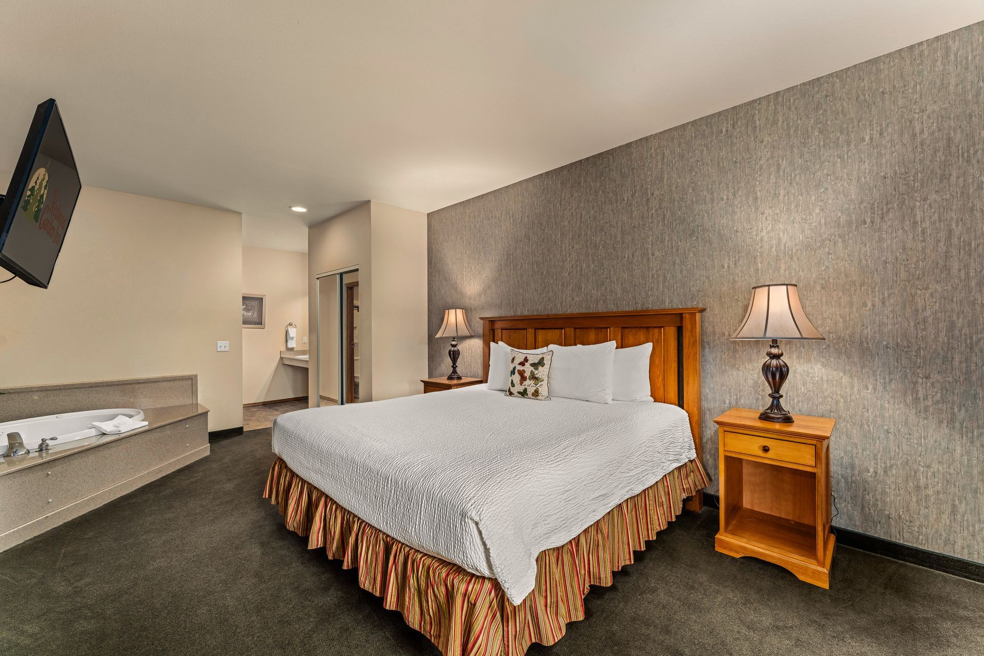 Hotel room with queen bed, bedside table, wall TV, and bench seating on a gray carpeted floor