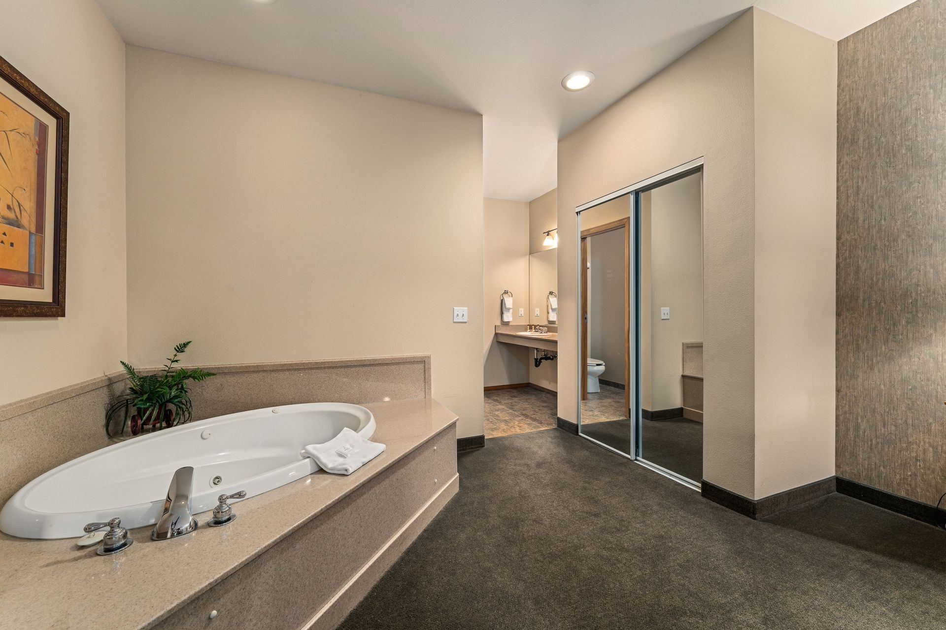 Modern bathroom with a large corner tub, beige walls, and dark carpet flooring