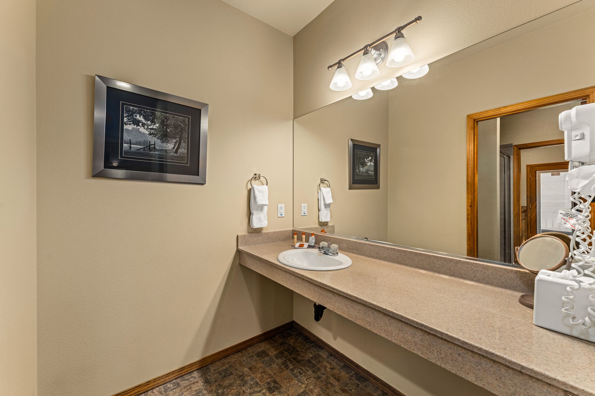 Hotel bathroom vanity with sink, beige walls, mirror, and overhead lights