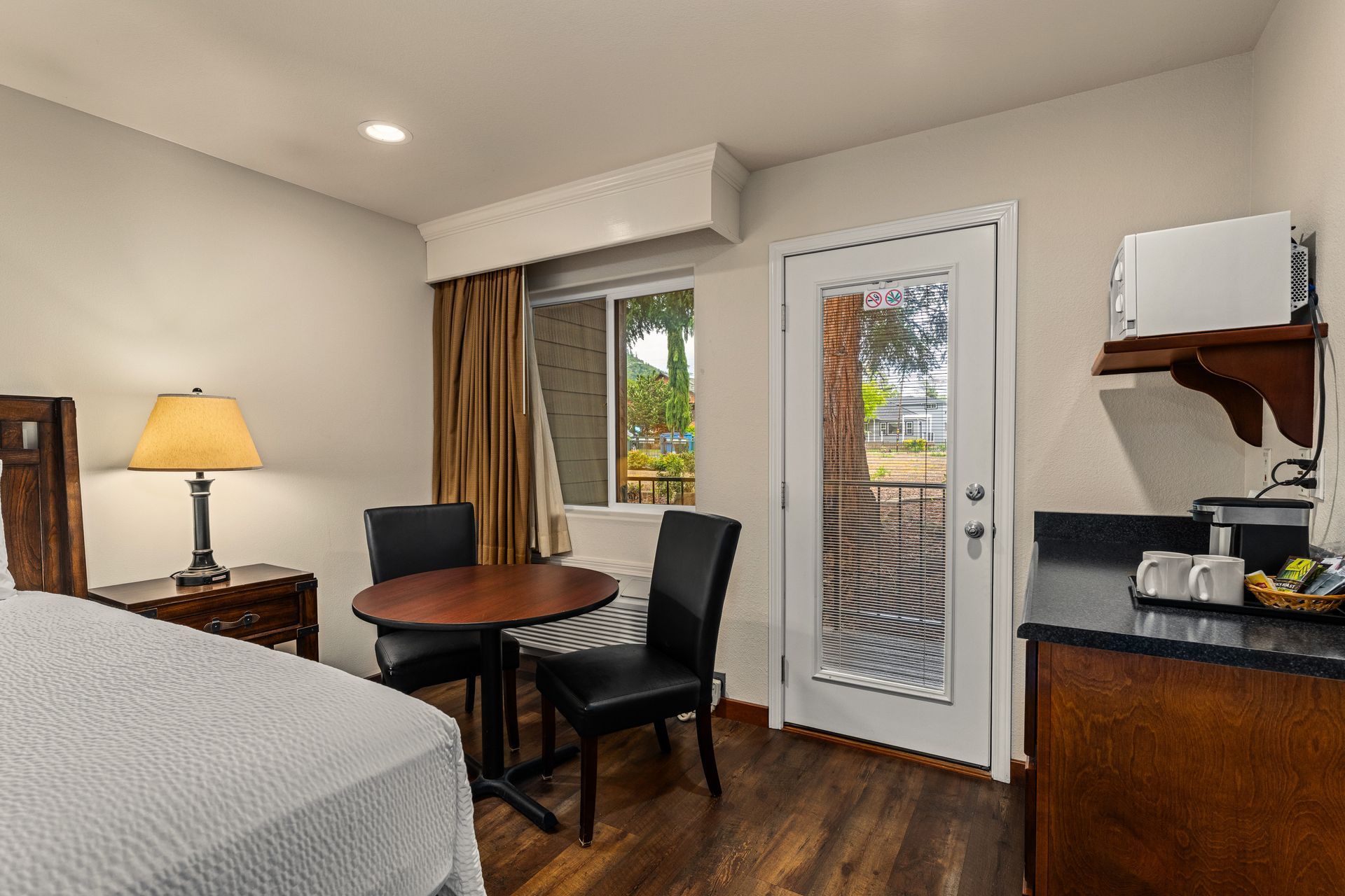 Small hotel room with bed, table and chairs, lamp, mini fridge, and glass door to balcony.
