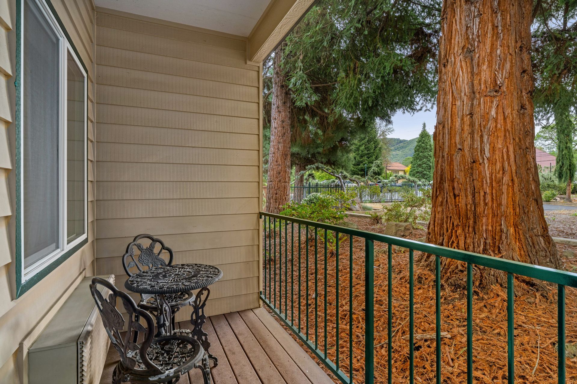 Covered porch with black metal chairs, wooden floor, railing, and large trees overlooking a yard.