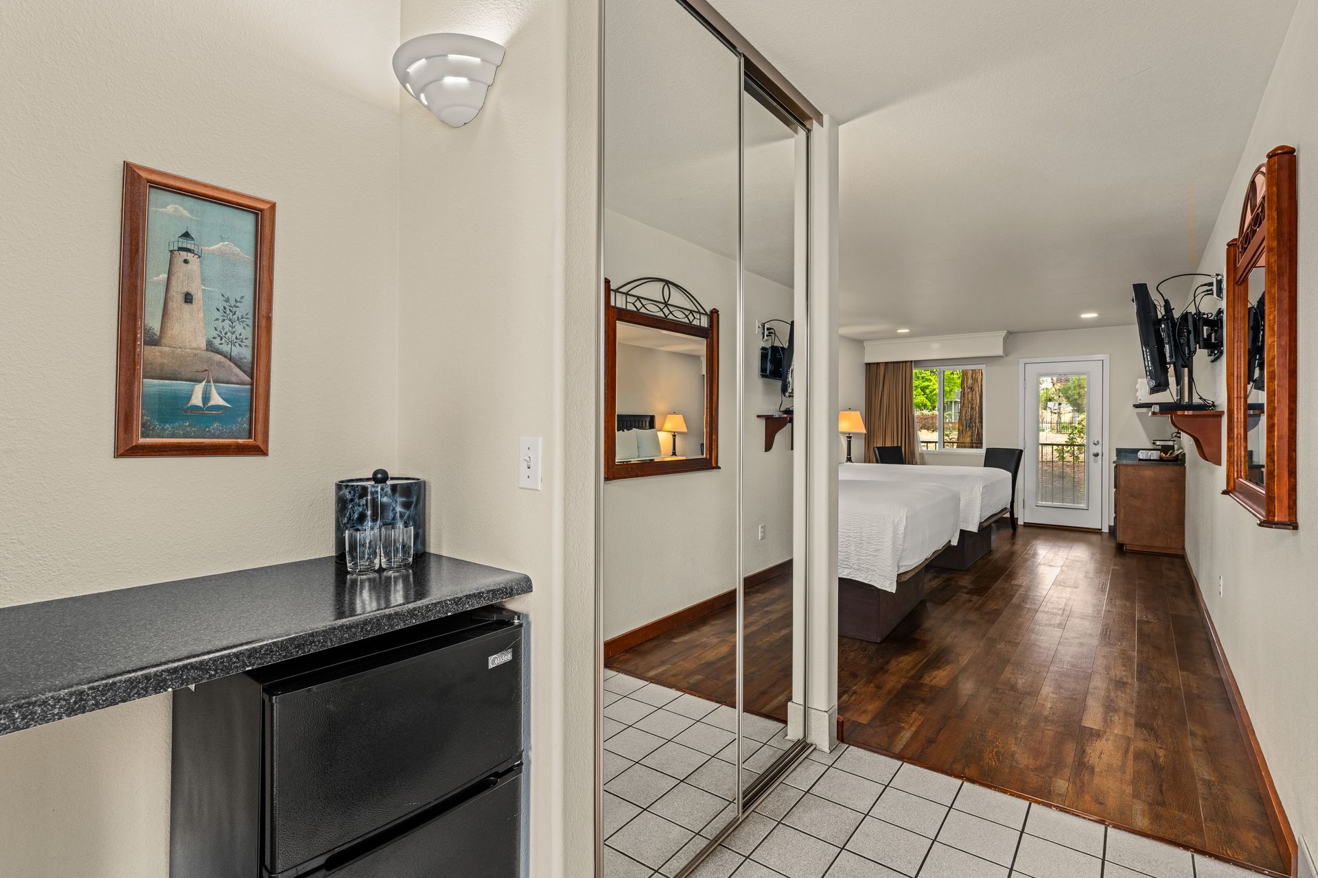 Small bedroom entryway with tiled floor, mirrored closet doors, a bed, and dark wood flooring leading to a window.