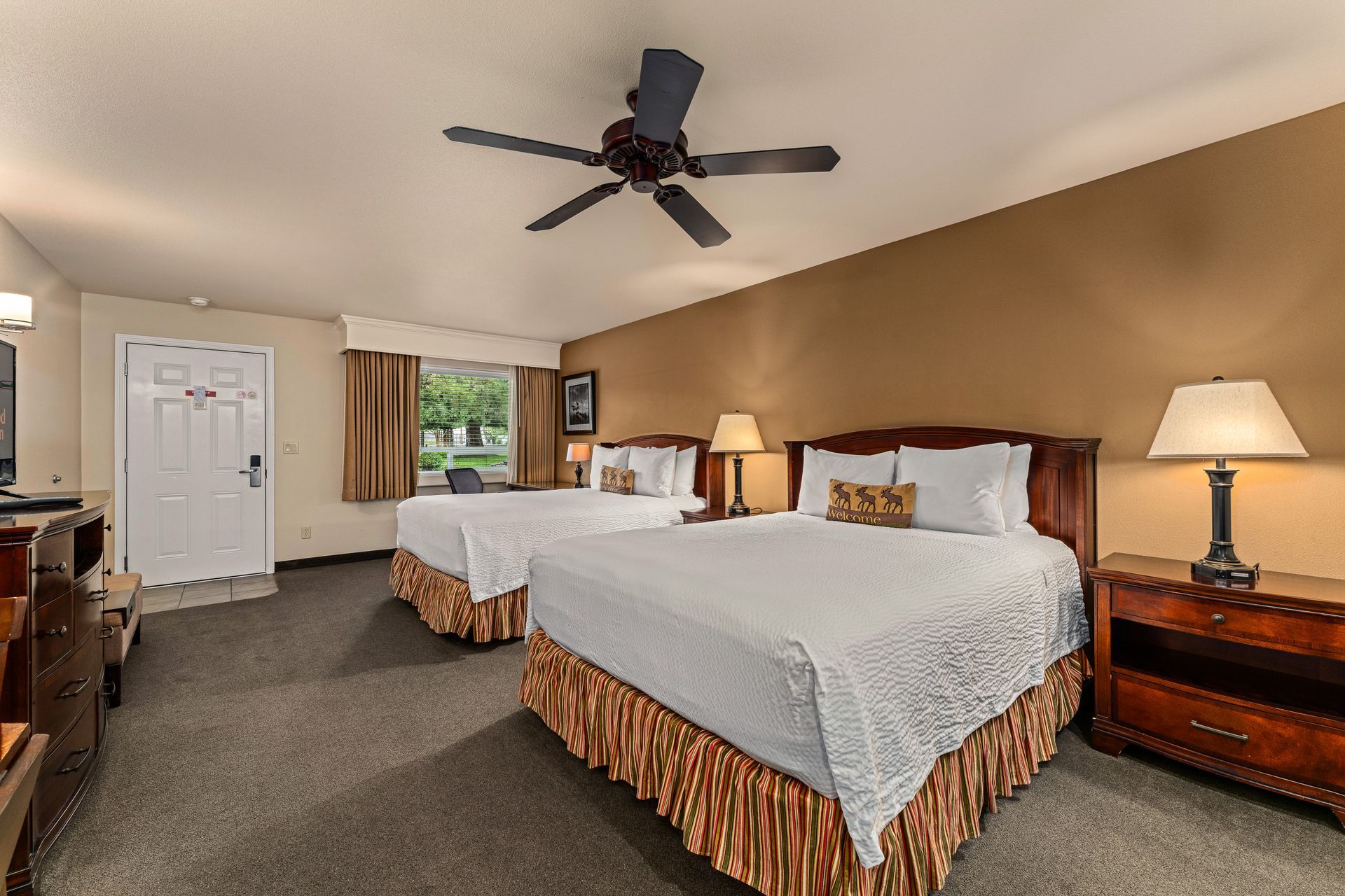 Hotel room with two queen beds, lamps, ceiling fan, and a window view.