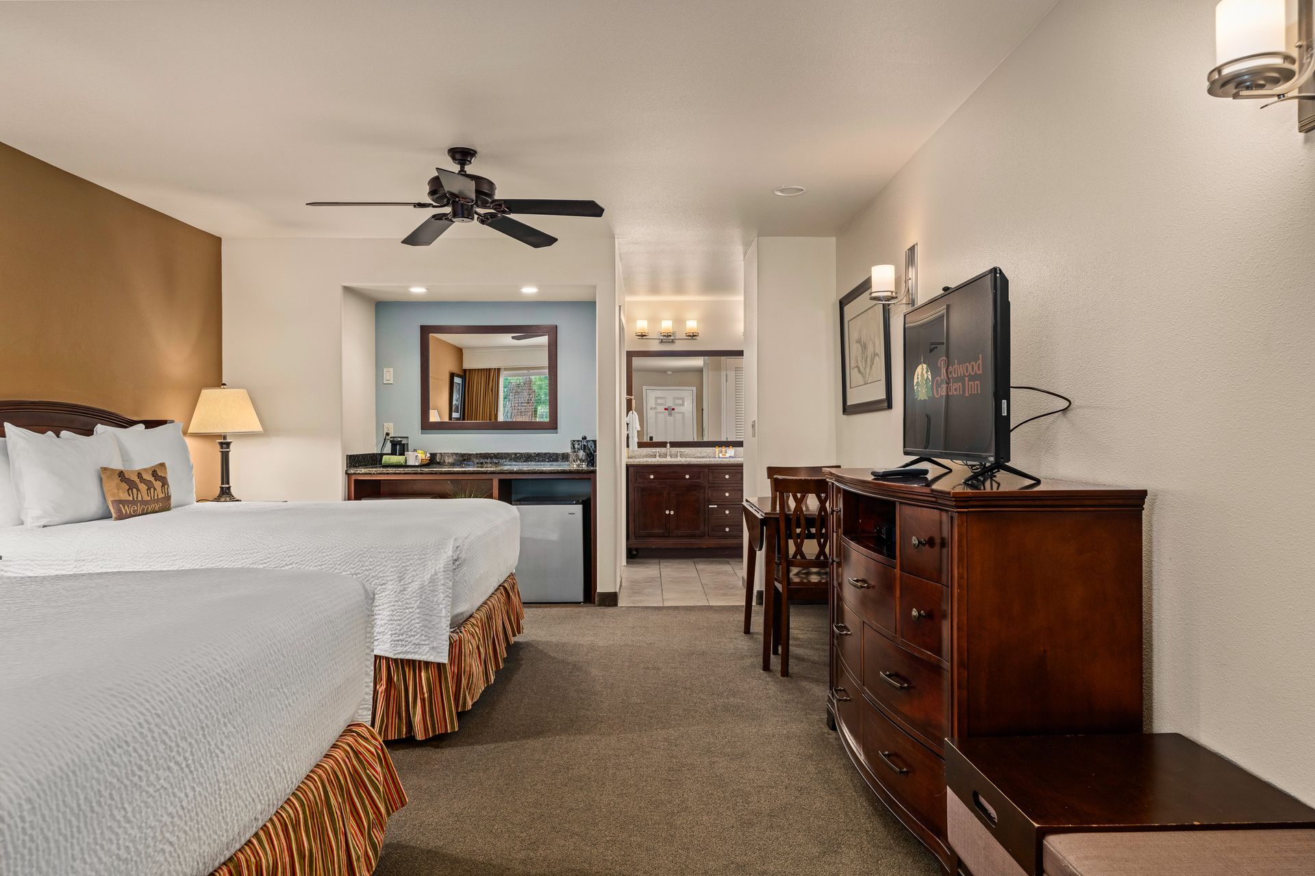 Hotel room with two beds, TV, desk, and ceiling fan in a beige interior