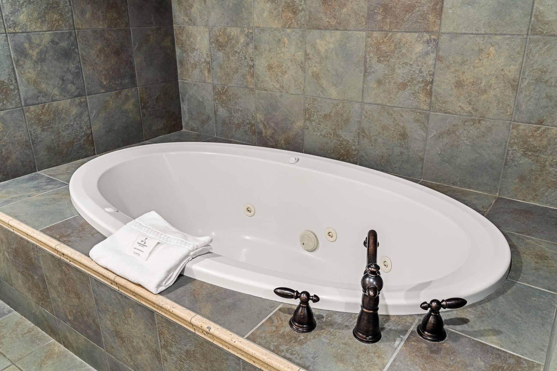 White oval bathtub with a folded towel, dark faucet fixtures, and tiled stone walls and floor