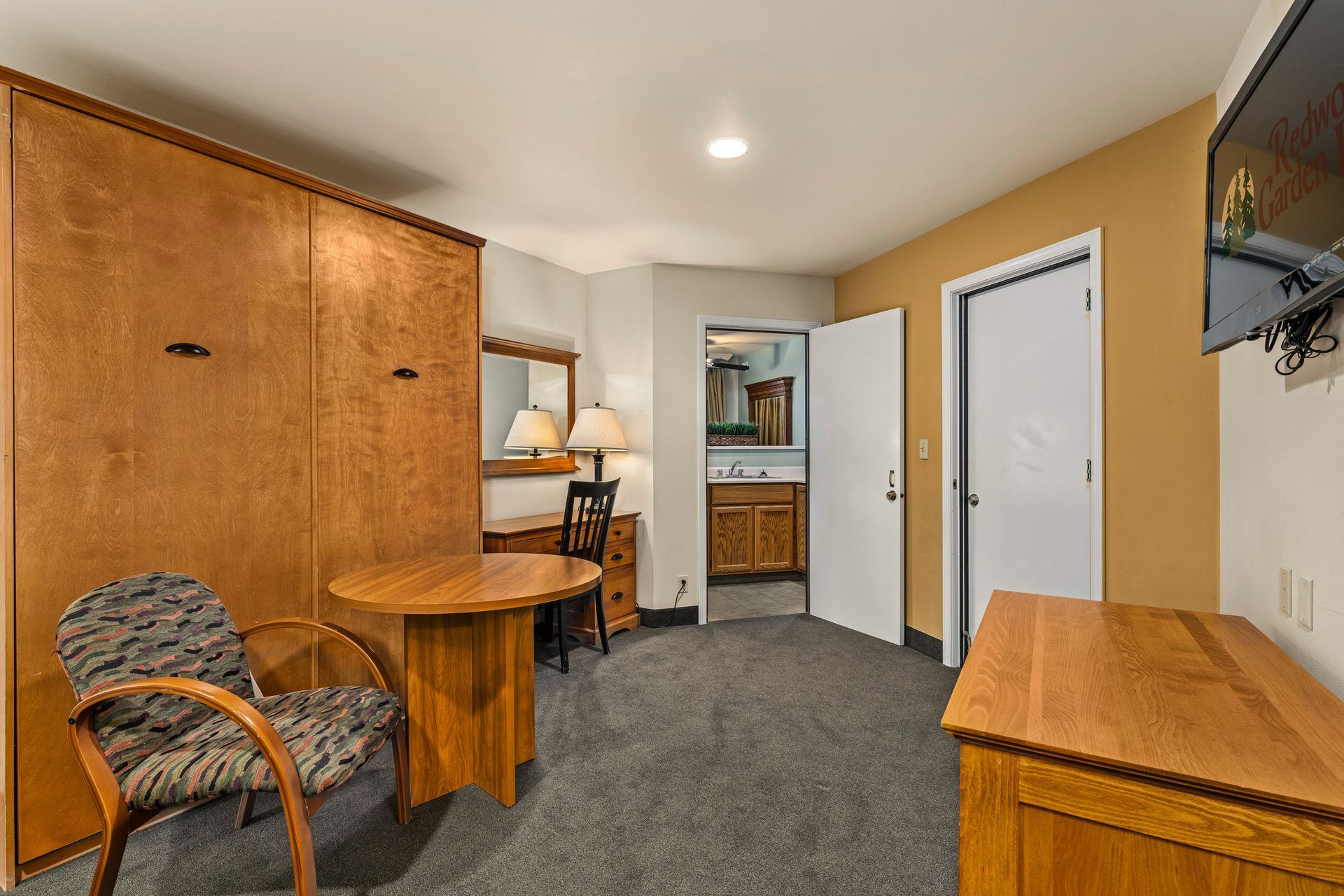 Small office with wood desks, a patterned chair, gray carpet, and an open doorway to a bathroom.