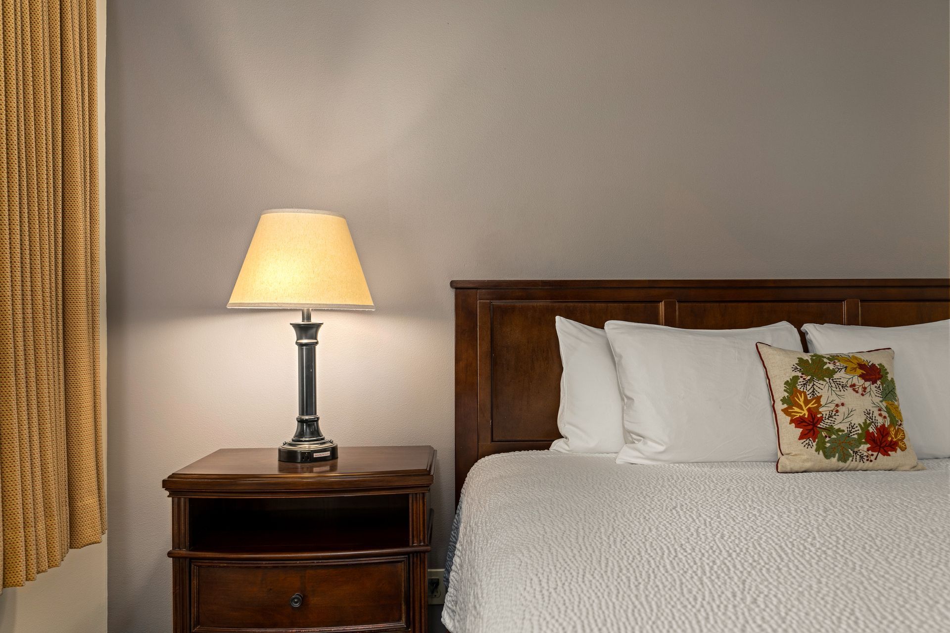 Bedroom with a lit lamp on a nightstand beside a bed with white bedding and a floral pillow.