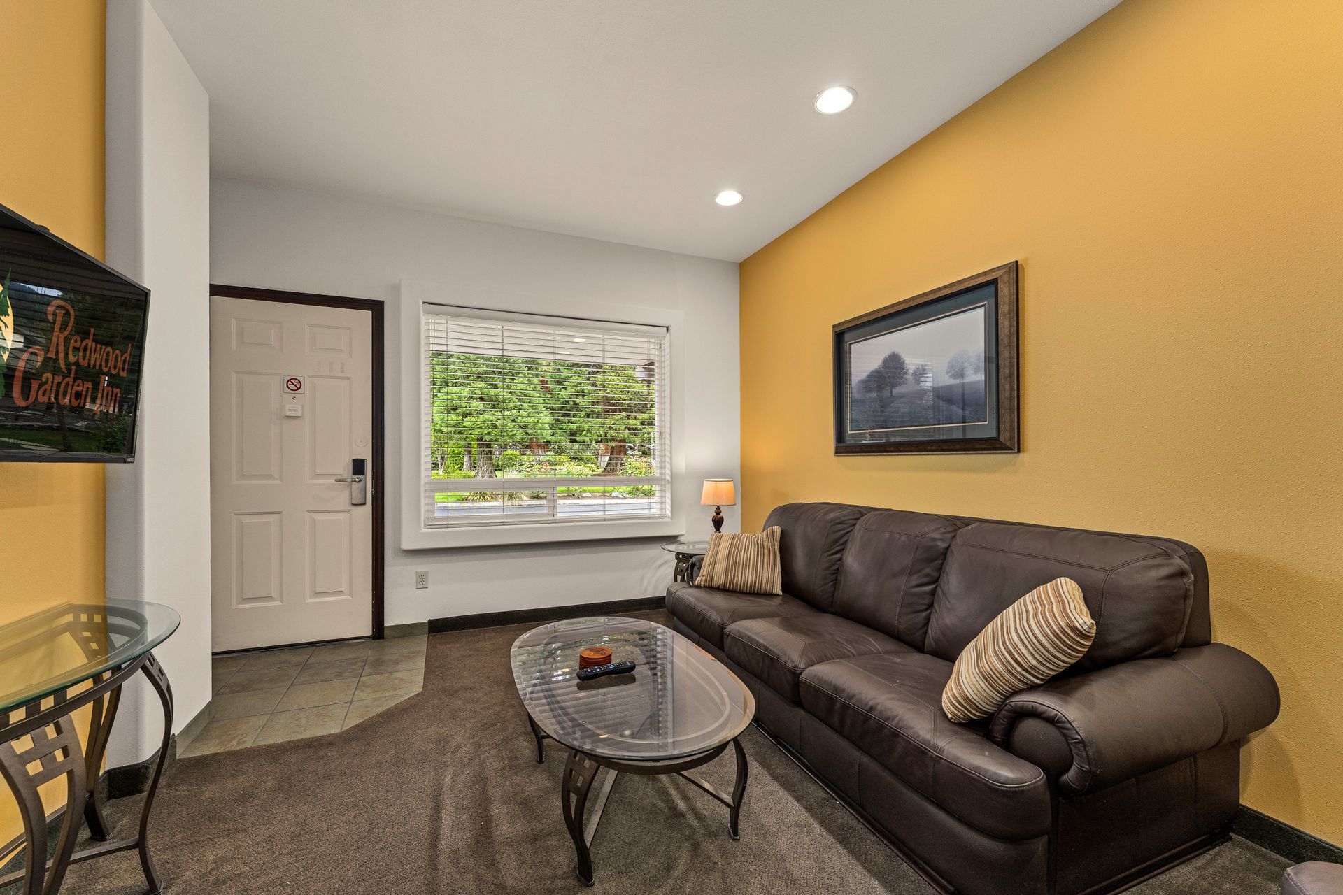 Cozy living room with brown sofa, glass coffee table, framed wall art, and window seat by a front door.