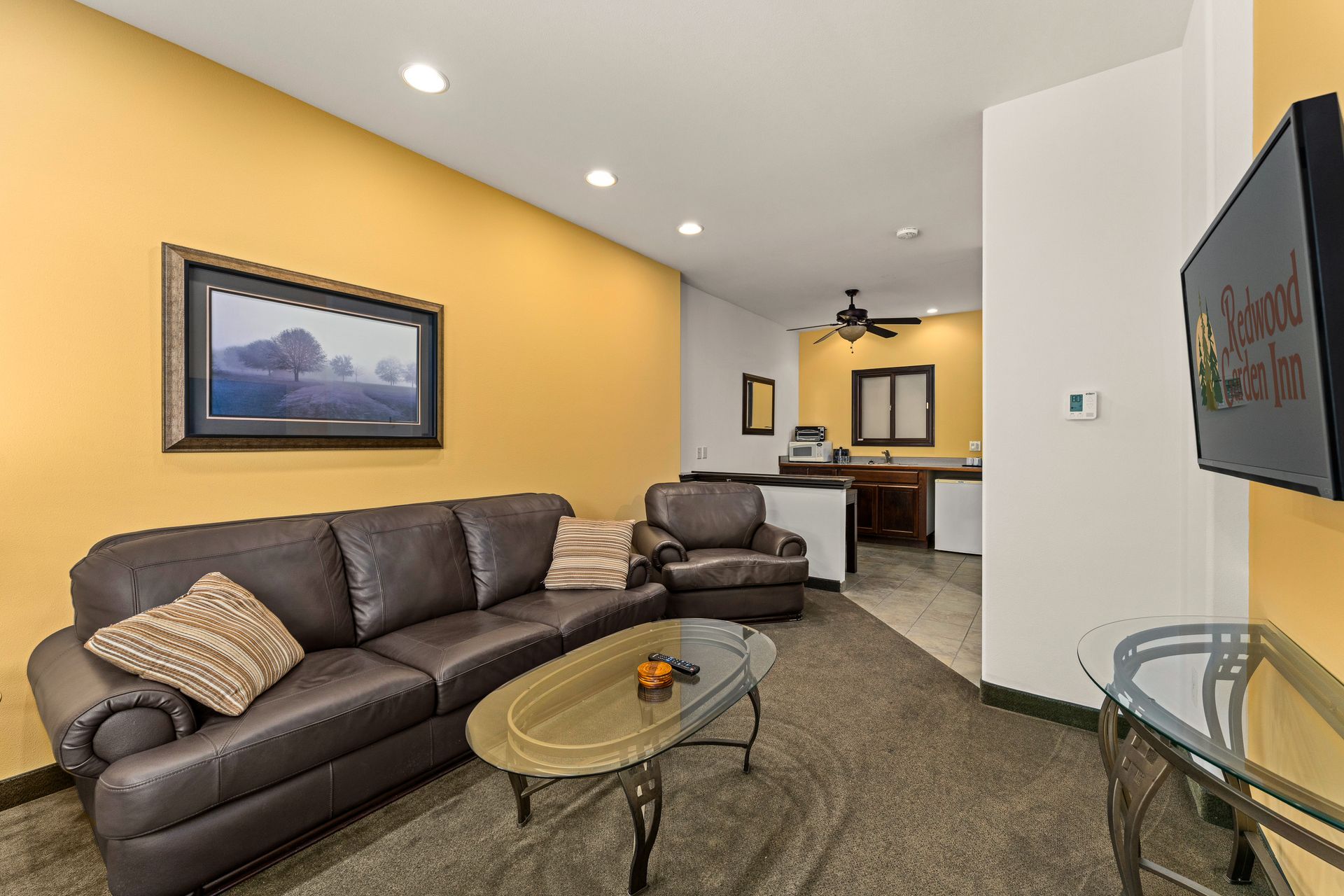 Cozy living room with brown leather sectional, glass coffee table, wall art, and open view to kitchen.
