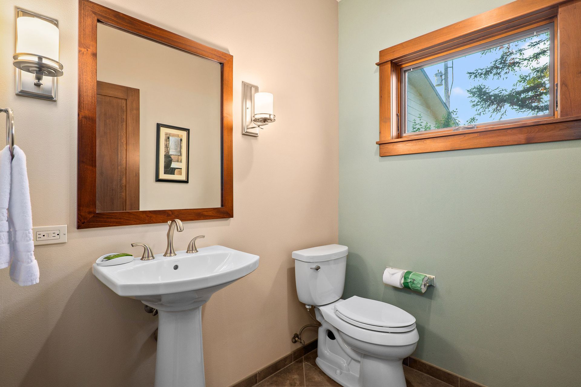 Small bathroom with pedestal sink, toilet, mirror, wall sconces, and a small window above the toilet.