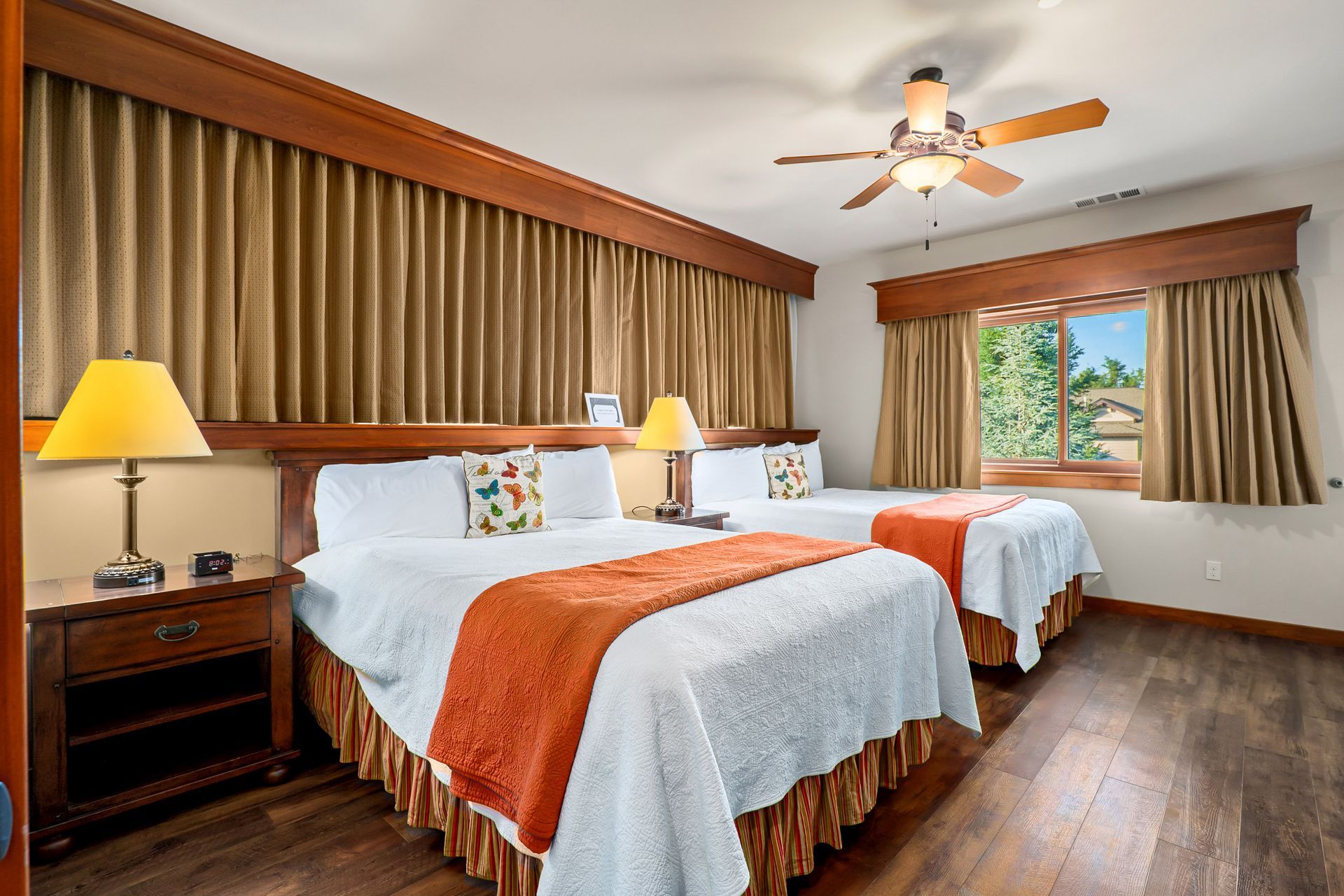 Hotel room with two white beds, orange accents, wooden floor, and a window with beige curtains