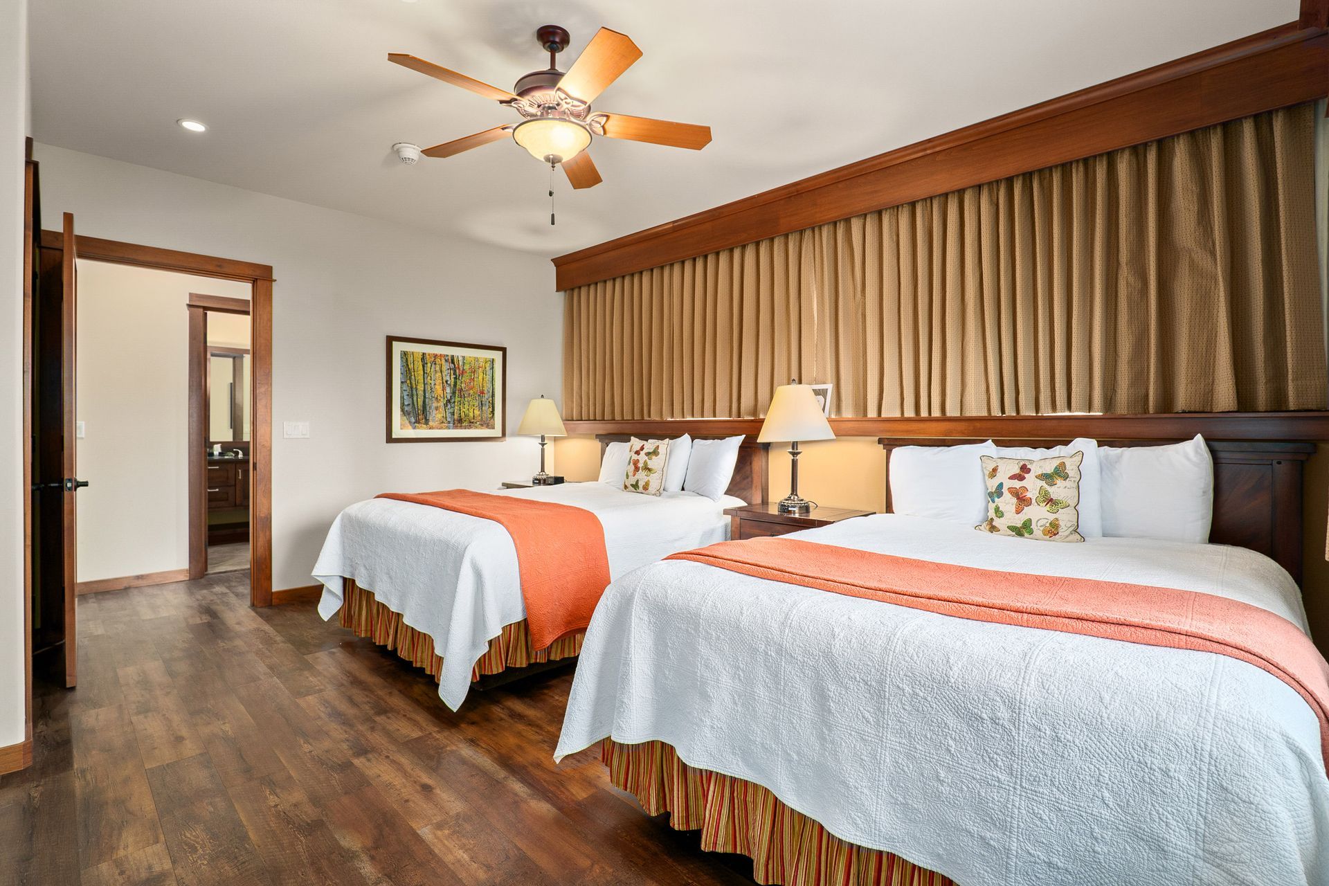 Hotel room with two queen beds, orange accents, wood floor, ceiling fan, and curtains