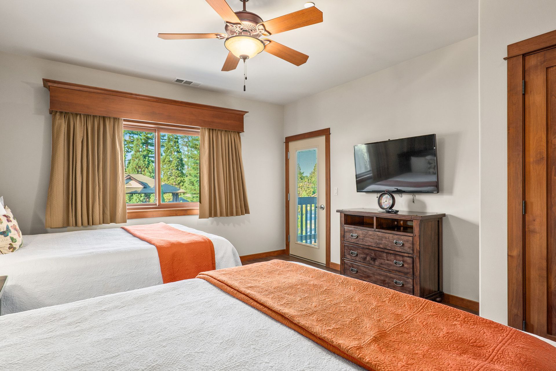 Bedroom with two beds, wood furniture, wall-mounted TV, ceiling fan, and a window with tan curtains