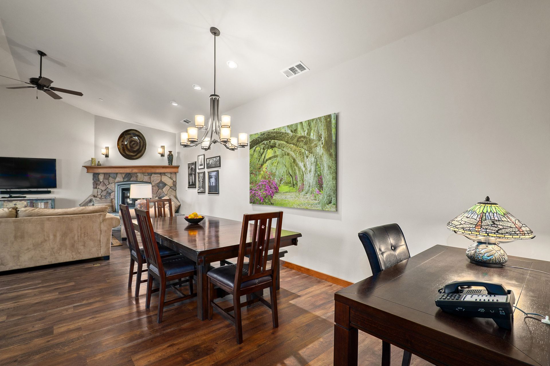 Dining room with dark wood table, chairs, chandelier, and framed green landscape art in an open home interior.