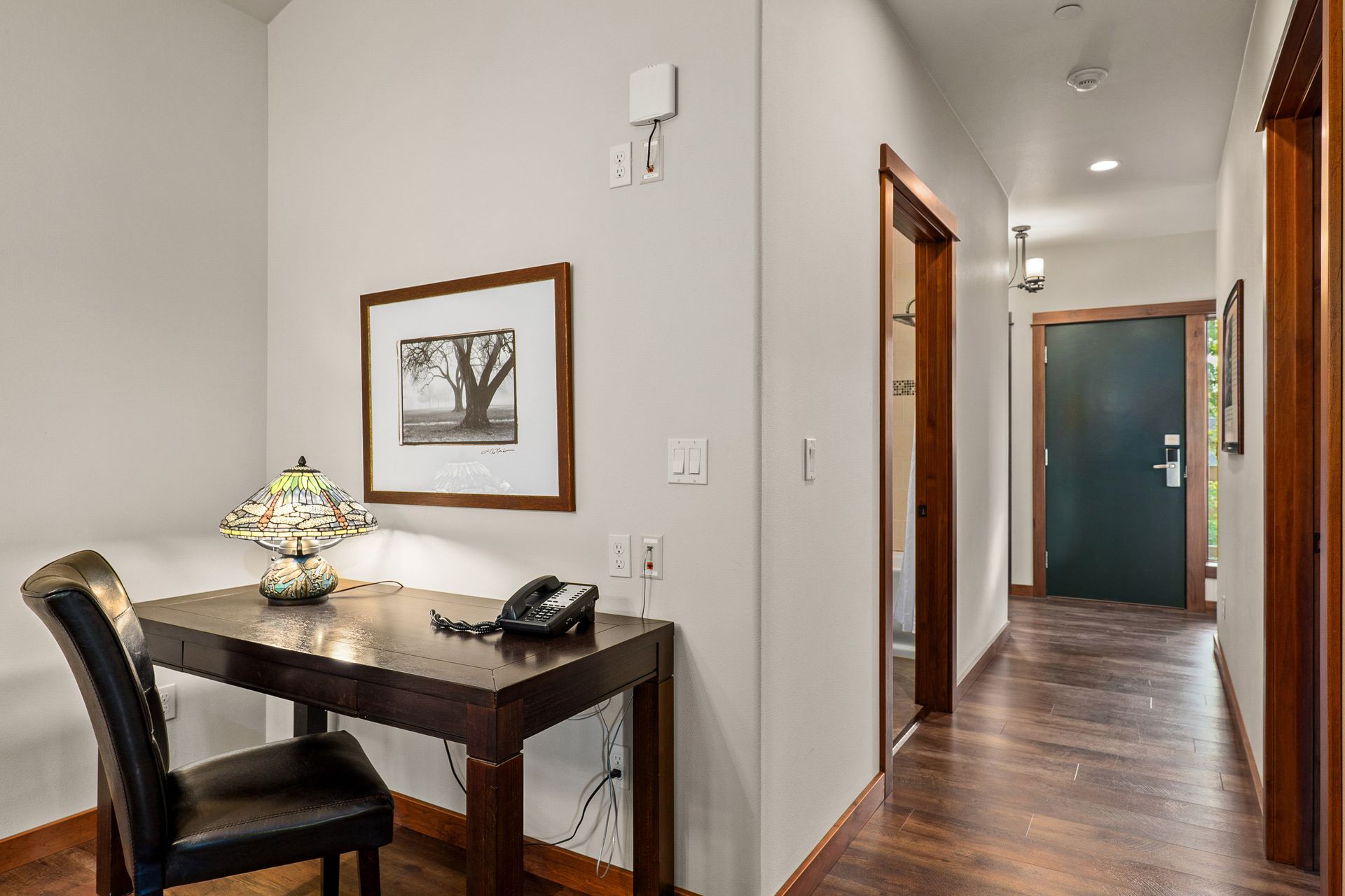 Hallway with dark wood floors, a small desk and chair, and framed wall art under a lit ceiling.