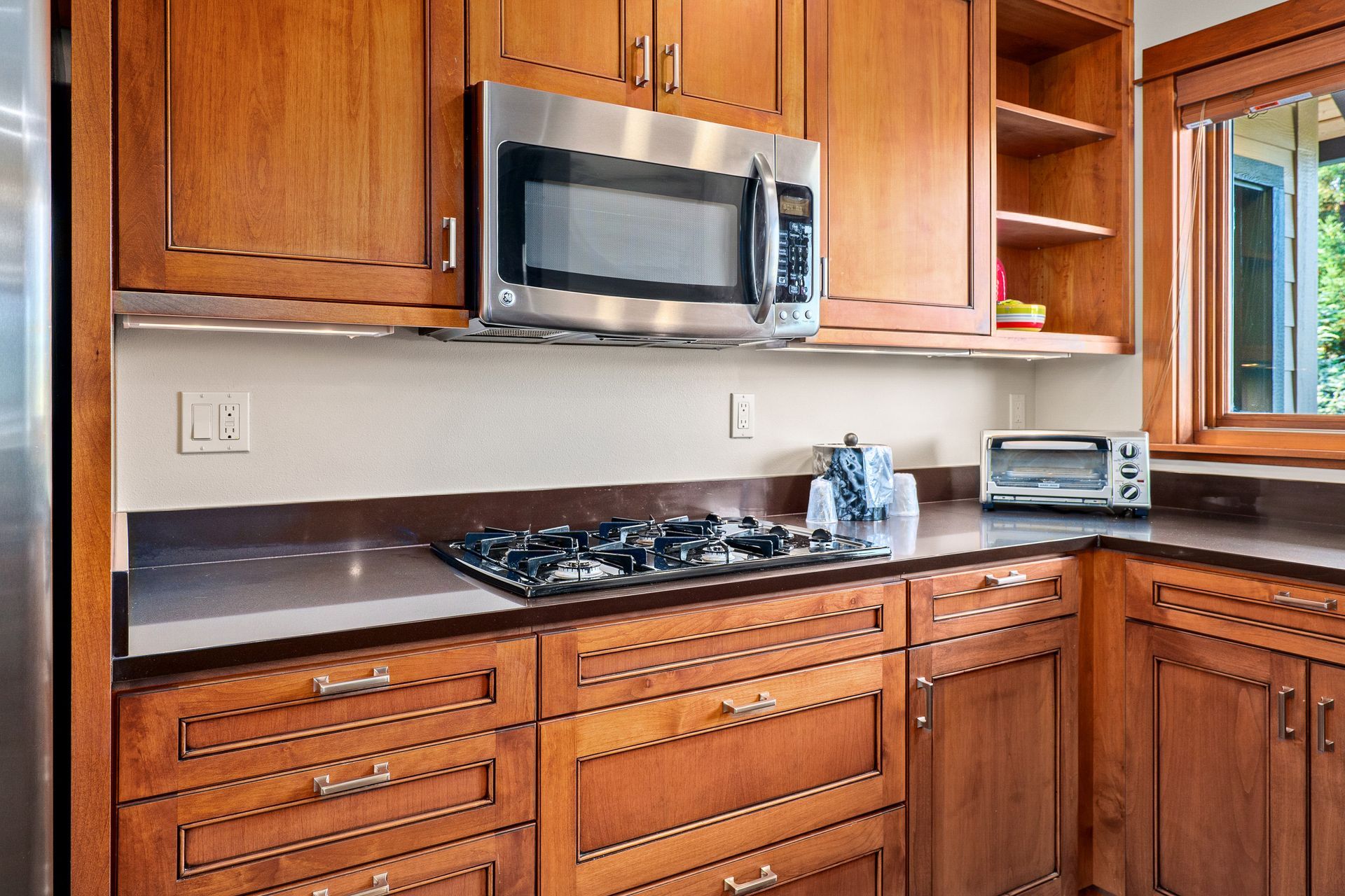 Wooden kitchen cabinets with stainless microwave, gas cooktop, and window by the sink