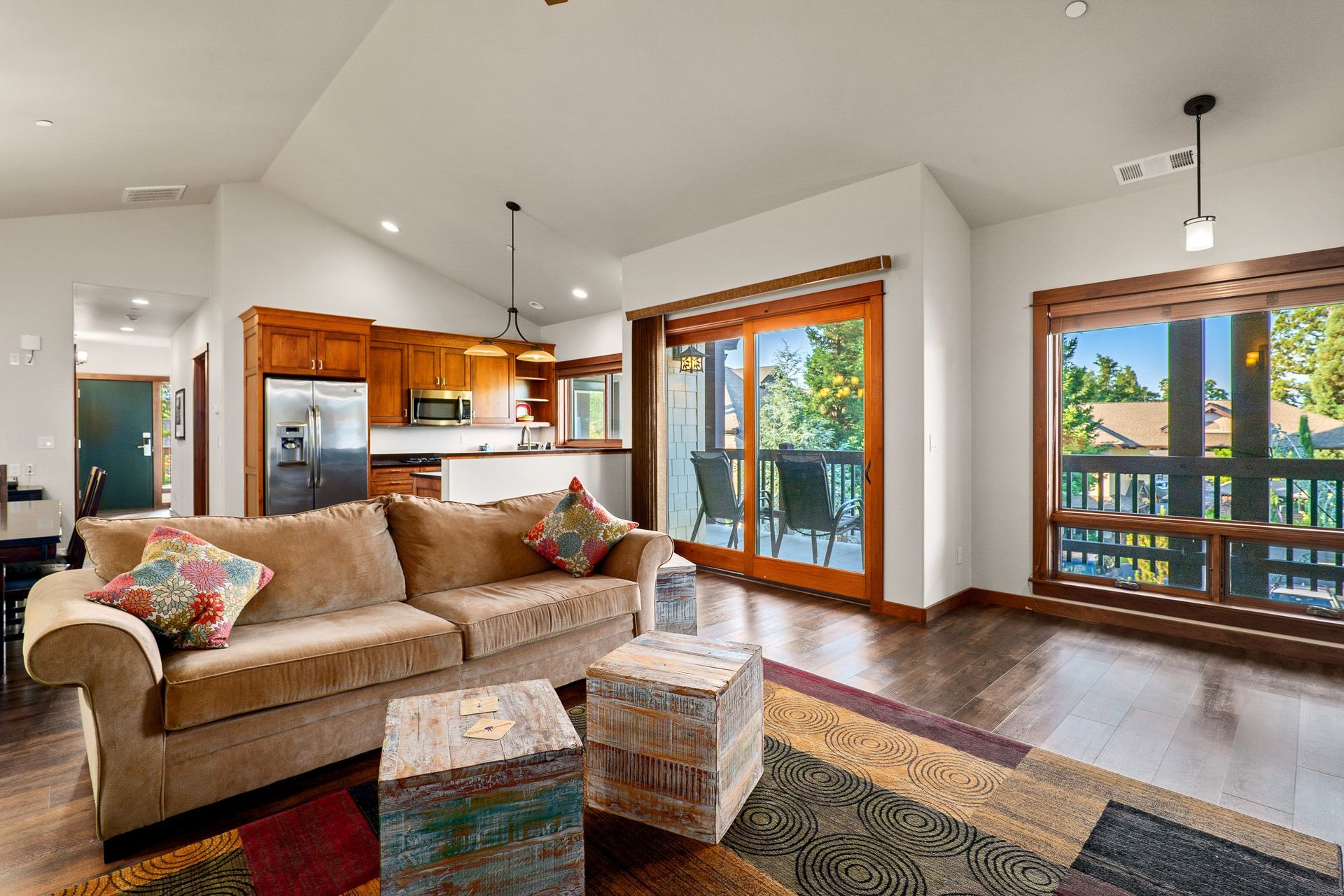Bright living room with tan sofa, colorful rug, kitchen, and large windows opening to a deck.