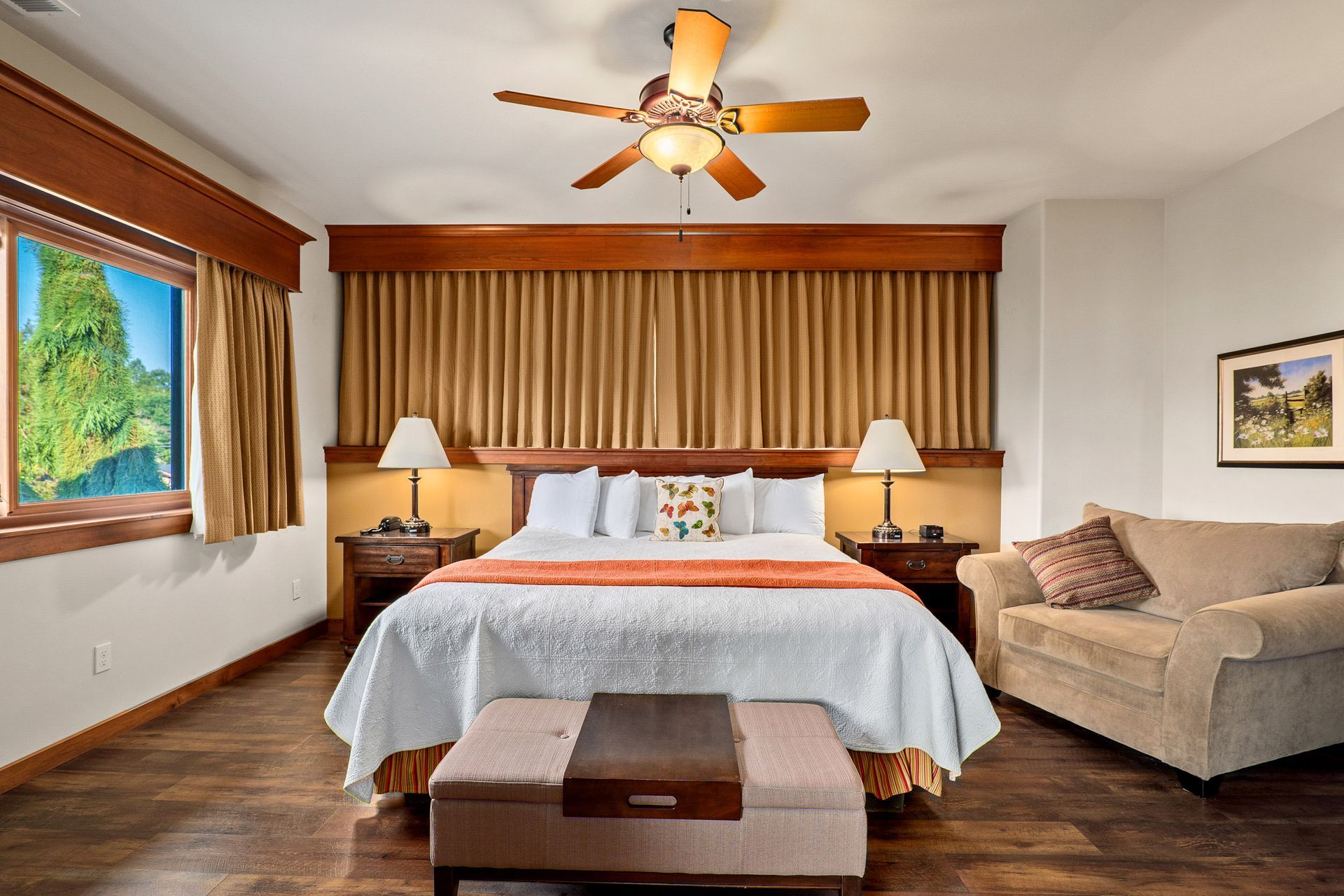 Cozy bedroom with a bed, beige sofa, wood floors, ceiling fan, and large window with mountain view