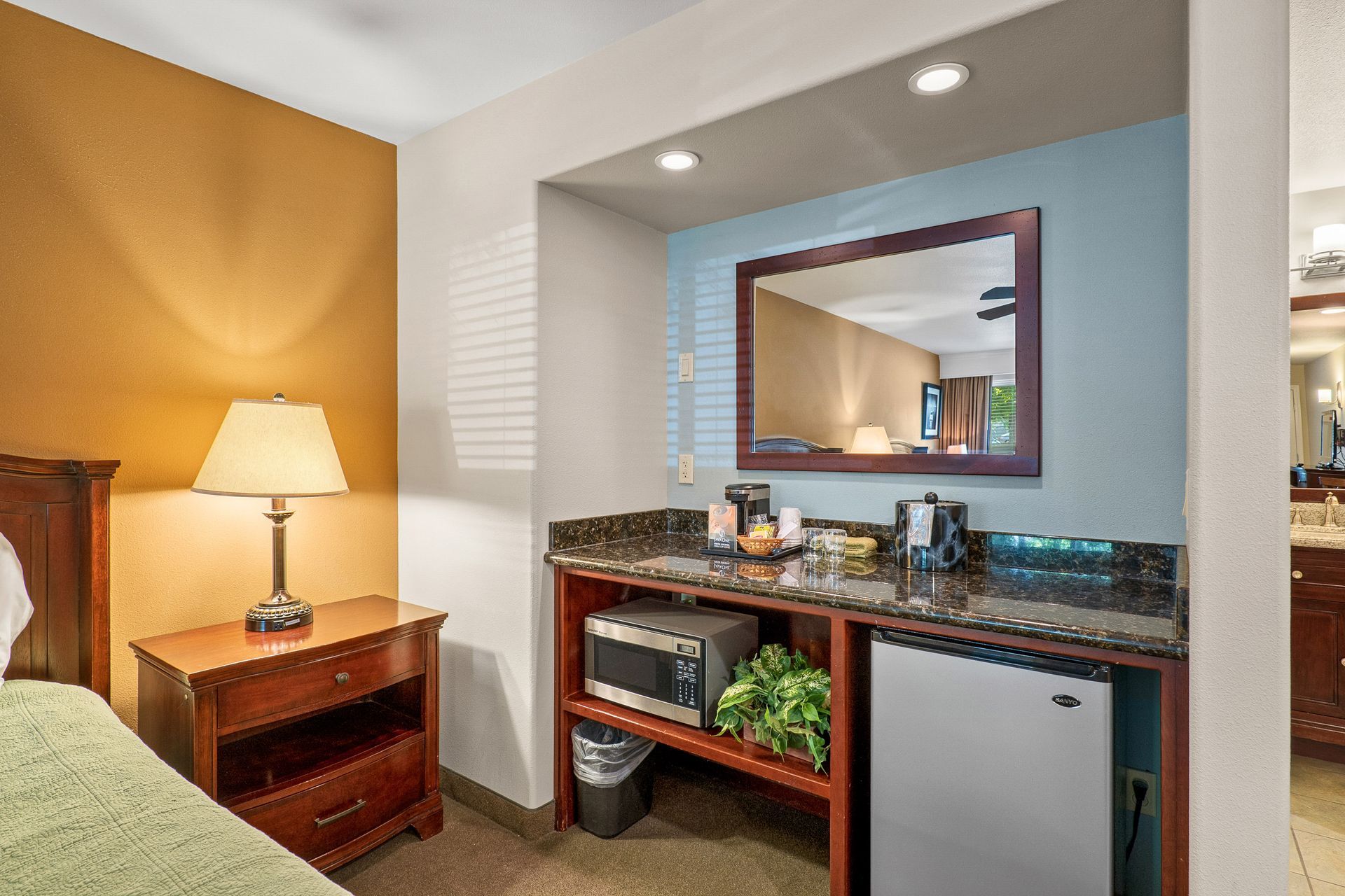 Hotel room vanity with mirror, sink, granite countertop, mini fridge, lamp, and bedside table