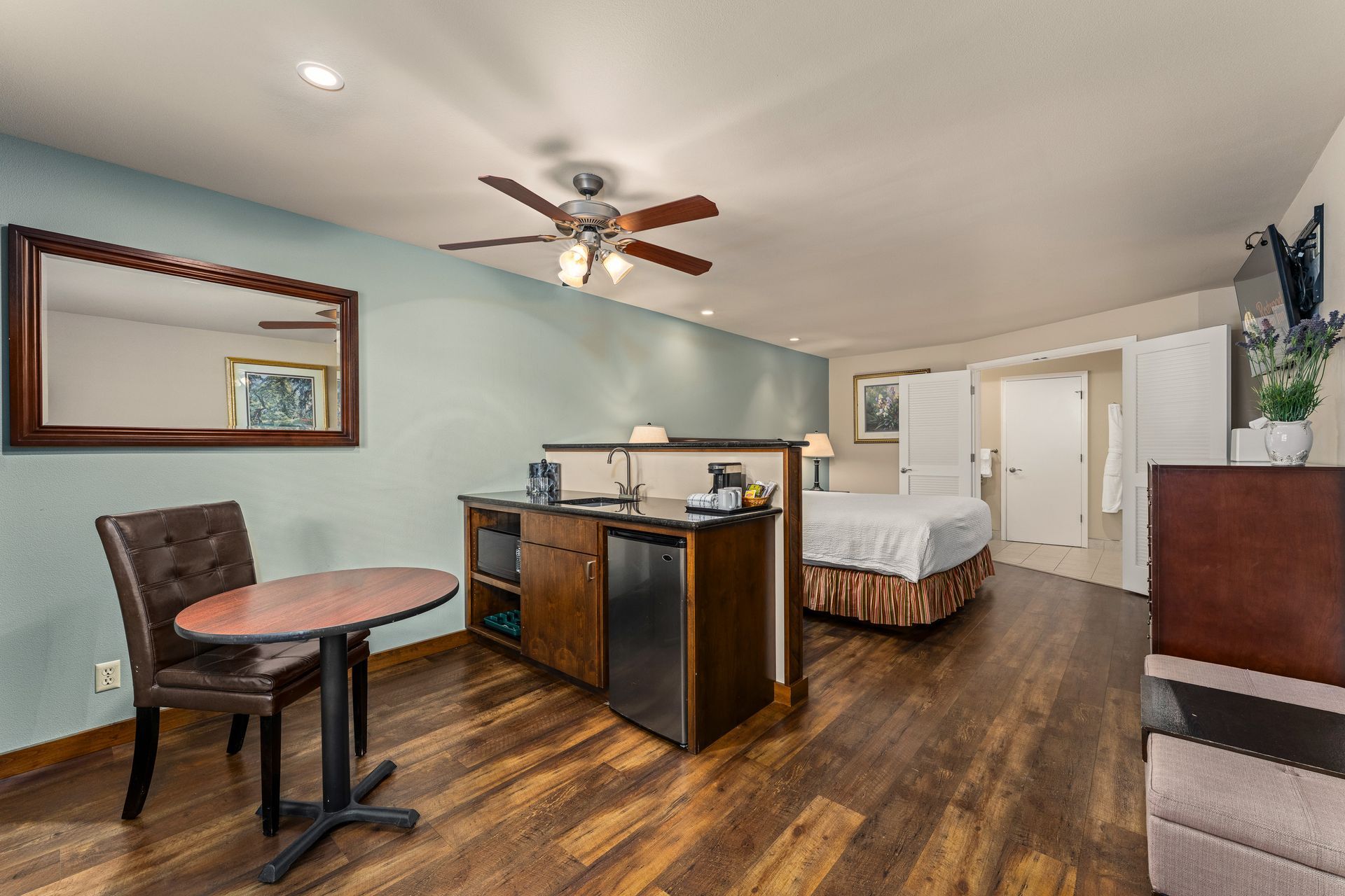 Spacious studio apartment with bed, dining table, desk, and wood flooring under a ceiling fan.