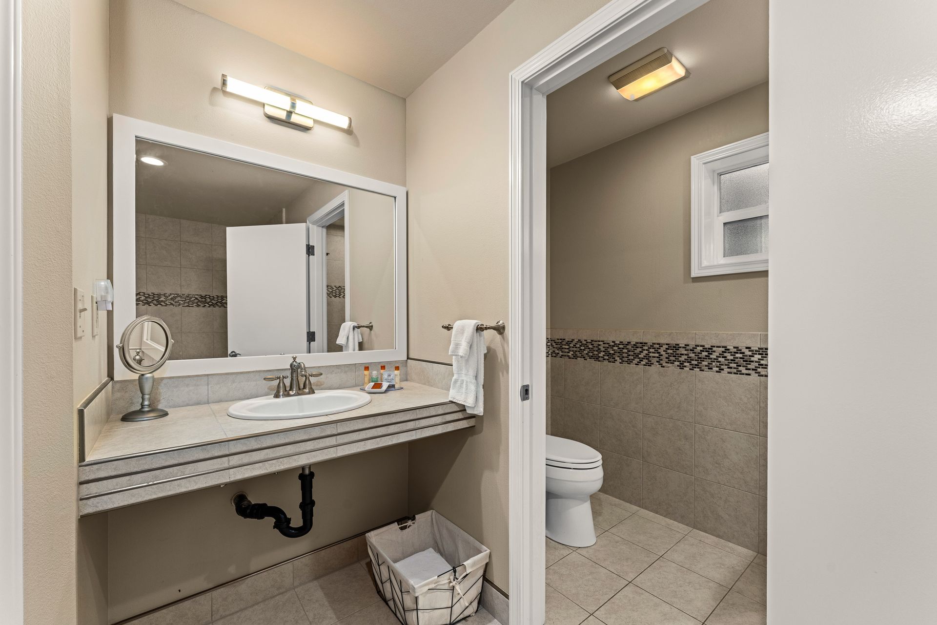 Bathroom with pedestal sink, large mirror, and open doorway to a toilet stall with beige tile.