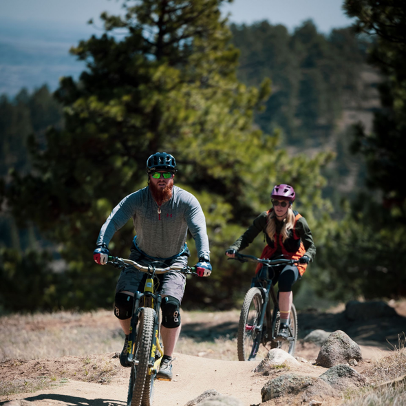 Two cyclists ride mountain bikes on a rocky dirt trail in a forested area.