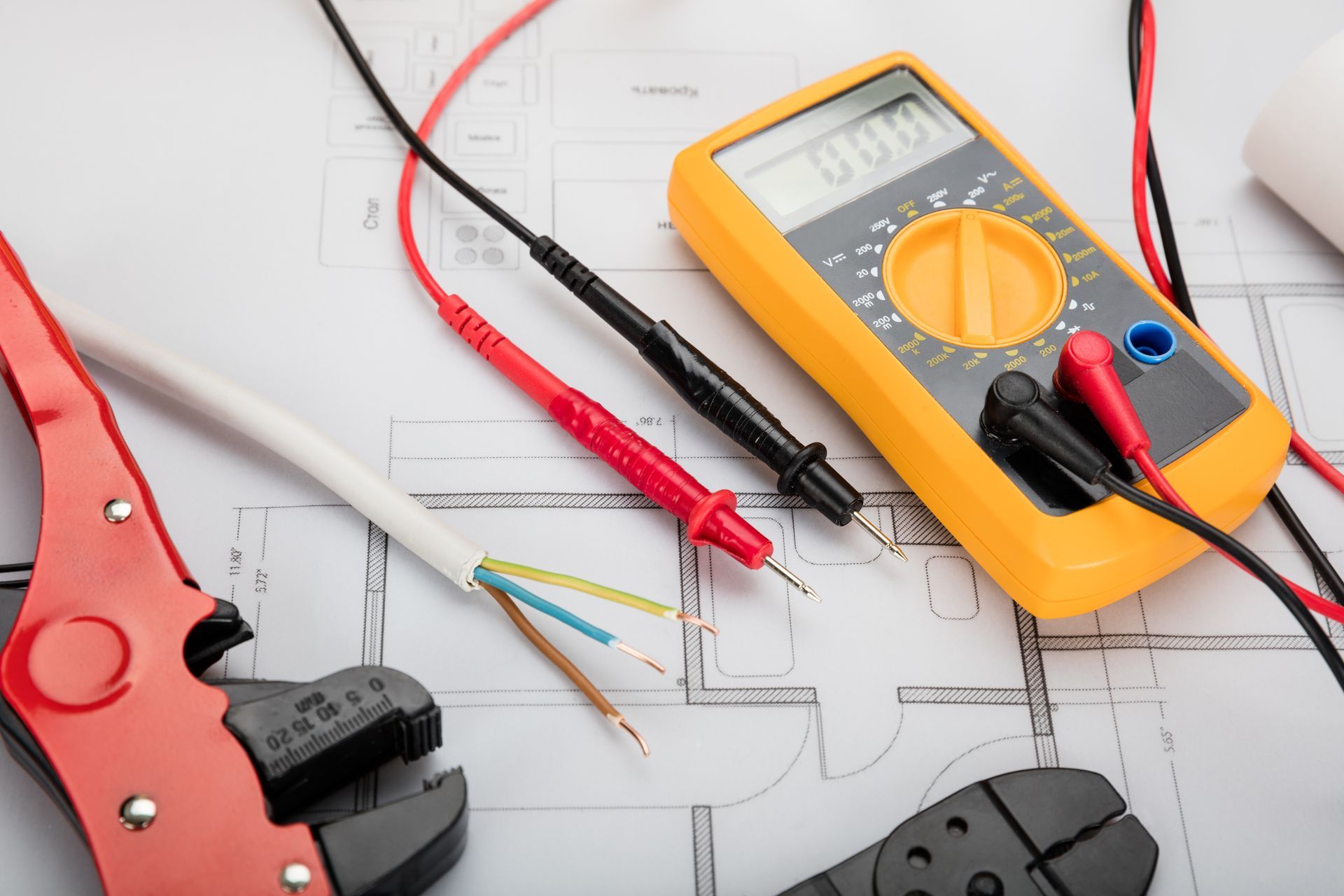 A yellow digital multimeter, red and black test leads, wire strippers, and an electrical cable on a floor plan blueprint.