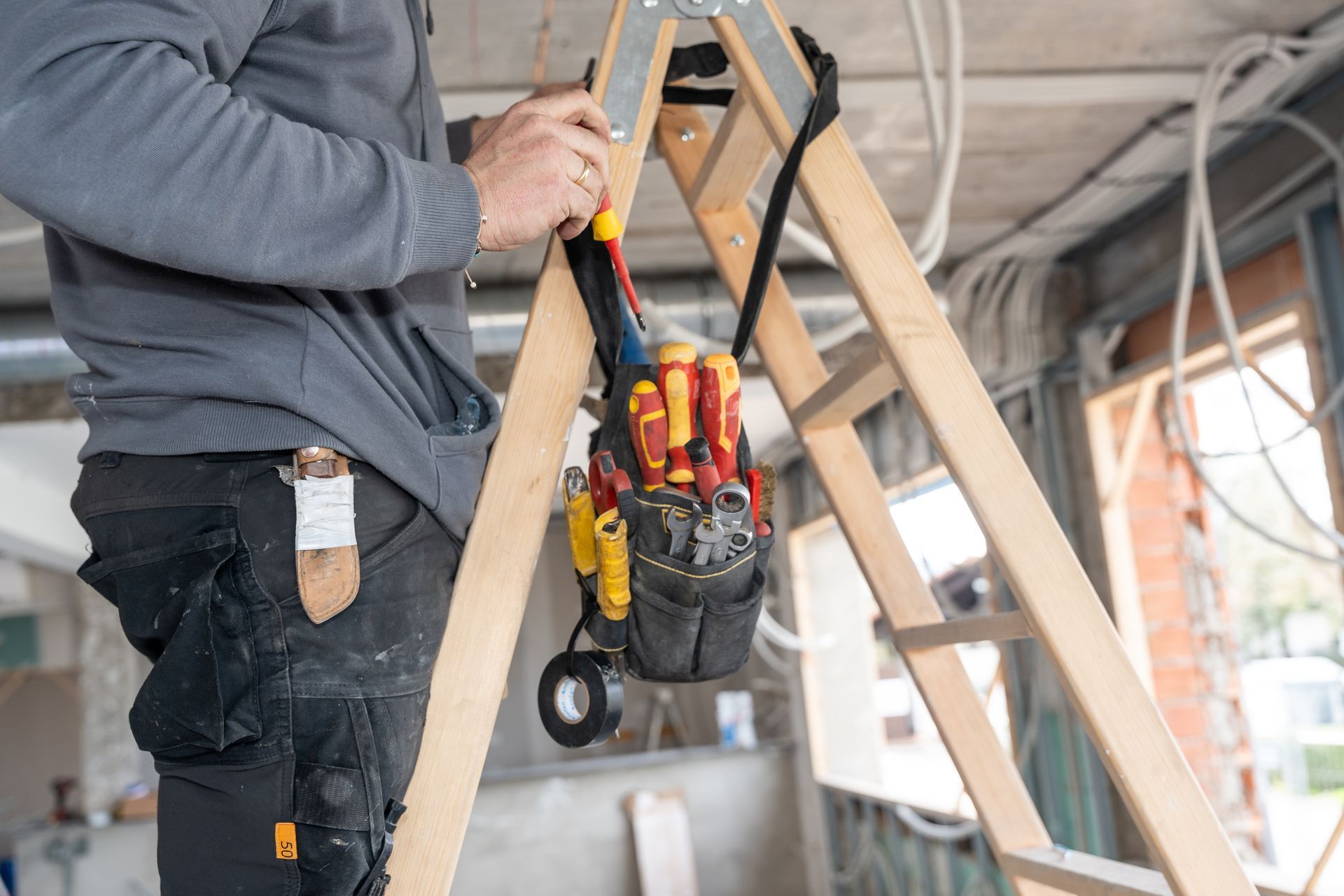Worker on a ladder carrying tools in a construction site, wearing a gray hoodie and black work pants.