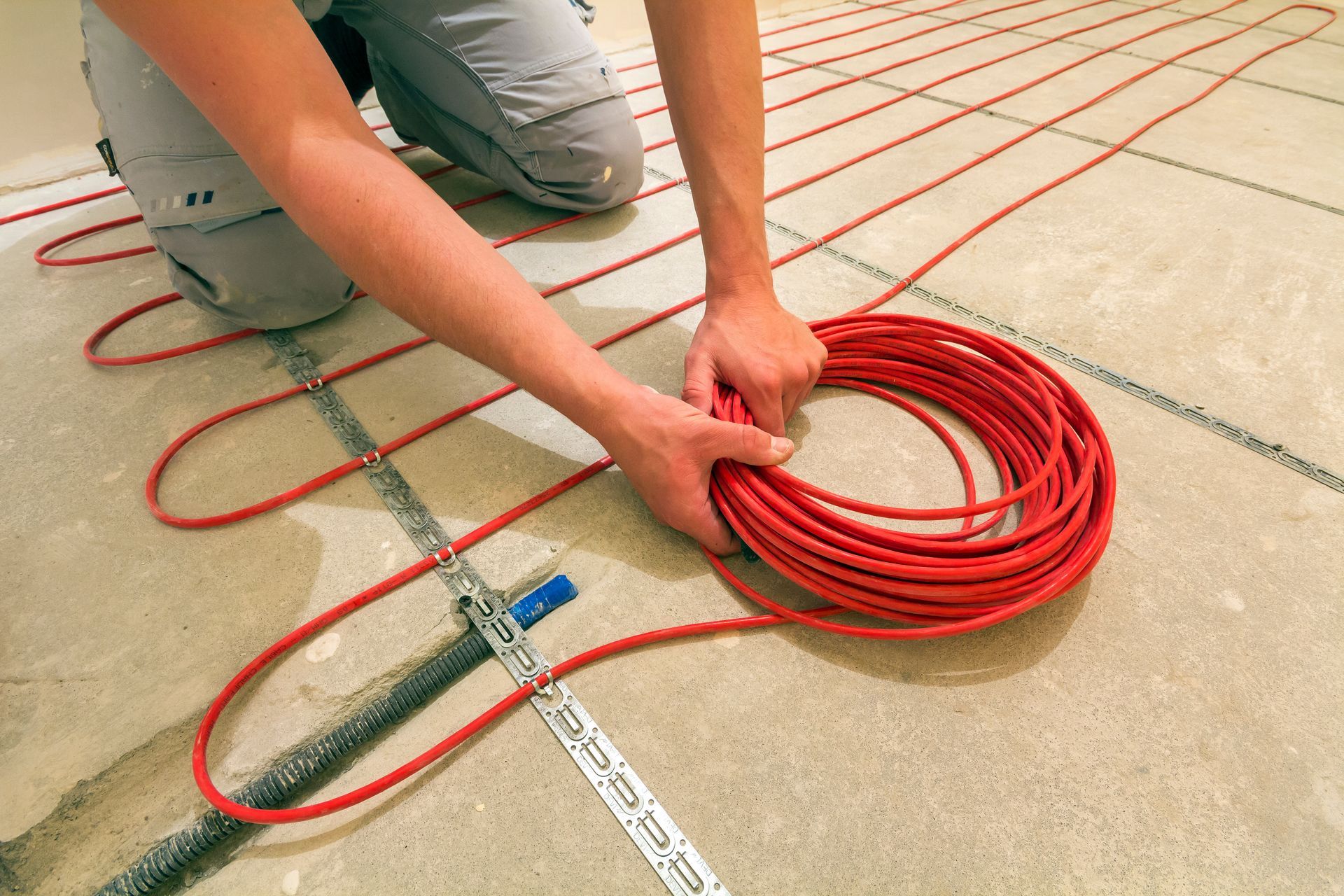 A worker kneeling on a floor, installing red radiant heating cable in a snake pattern using a metal mounting strip.
