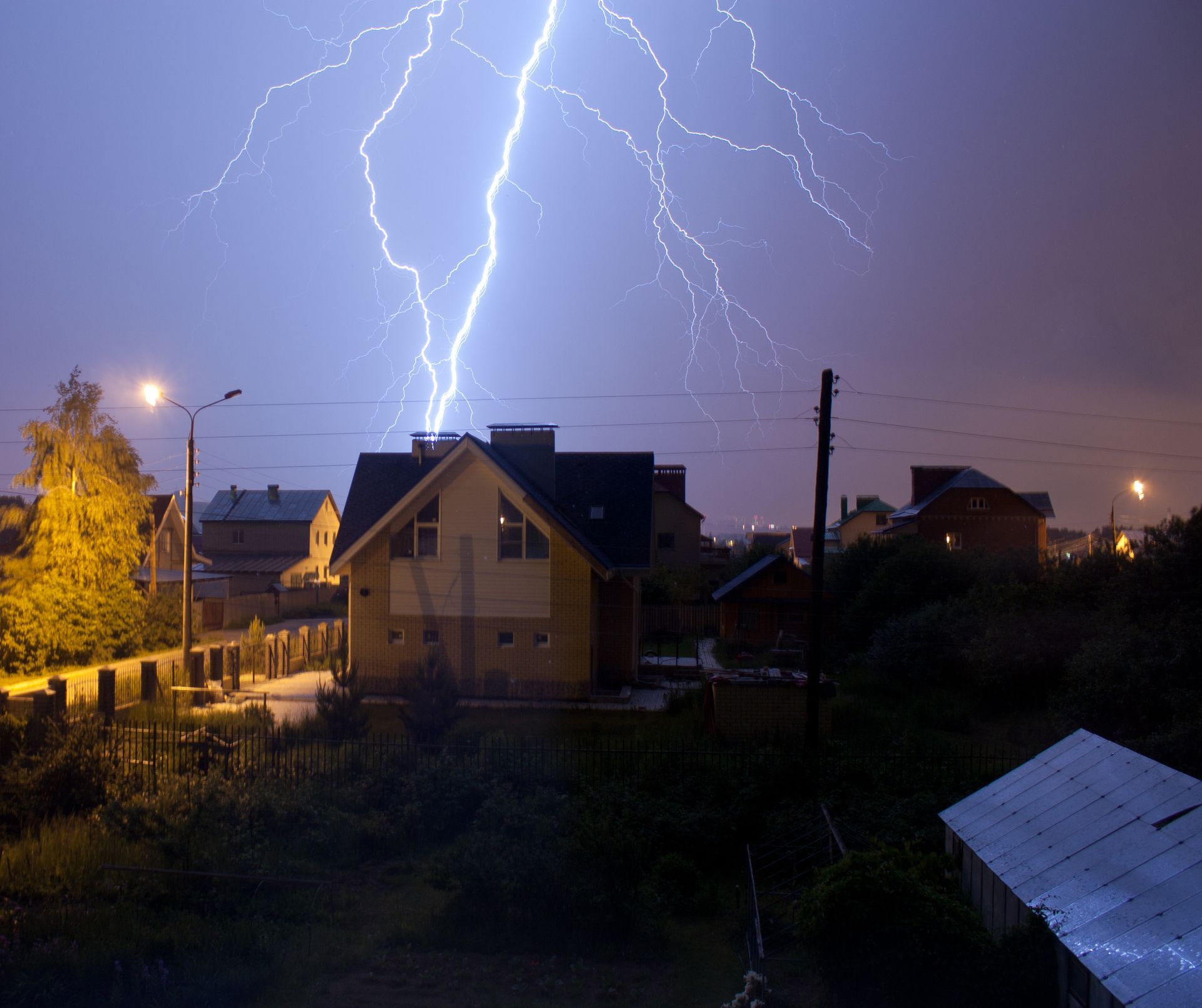 Lightning strikes over a neighborhood at night, illuminating houses and a streetlight.