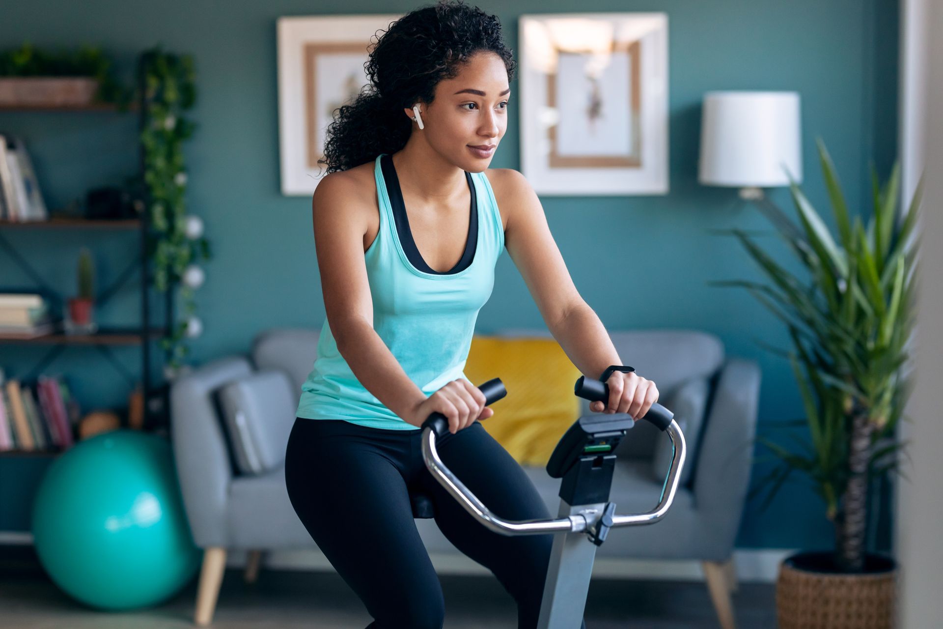 Woman exercising on a stationary bike in a bright living room with teal walls and a yoga ball.
