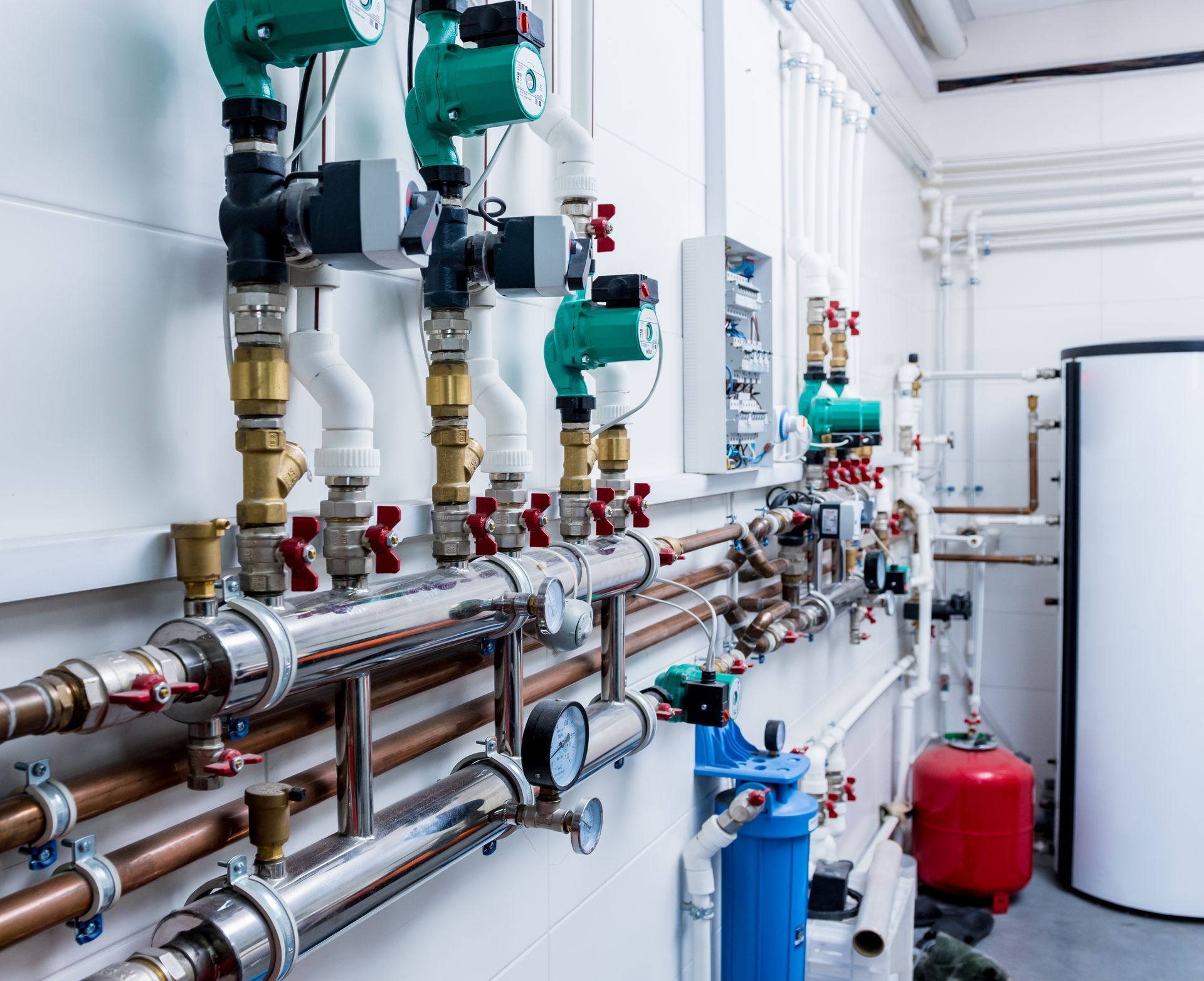 A wall-mounted industrial plumbing system featuring multiple green circulation pumps, stainless pipes, and gauges.
