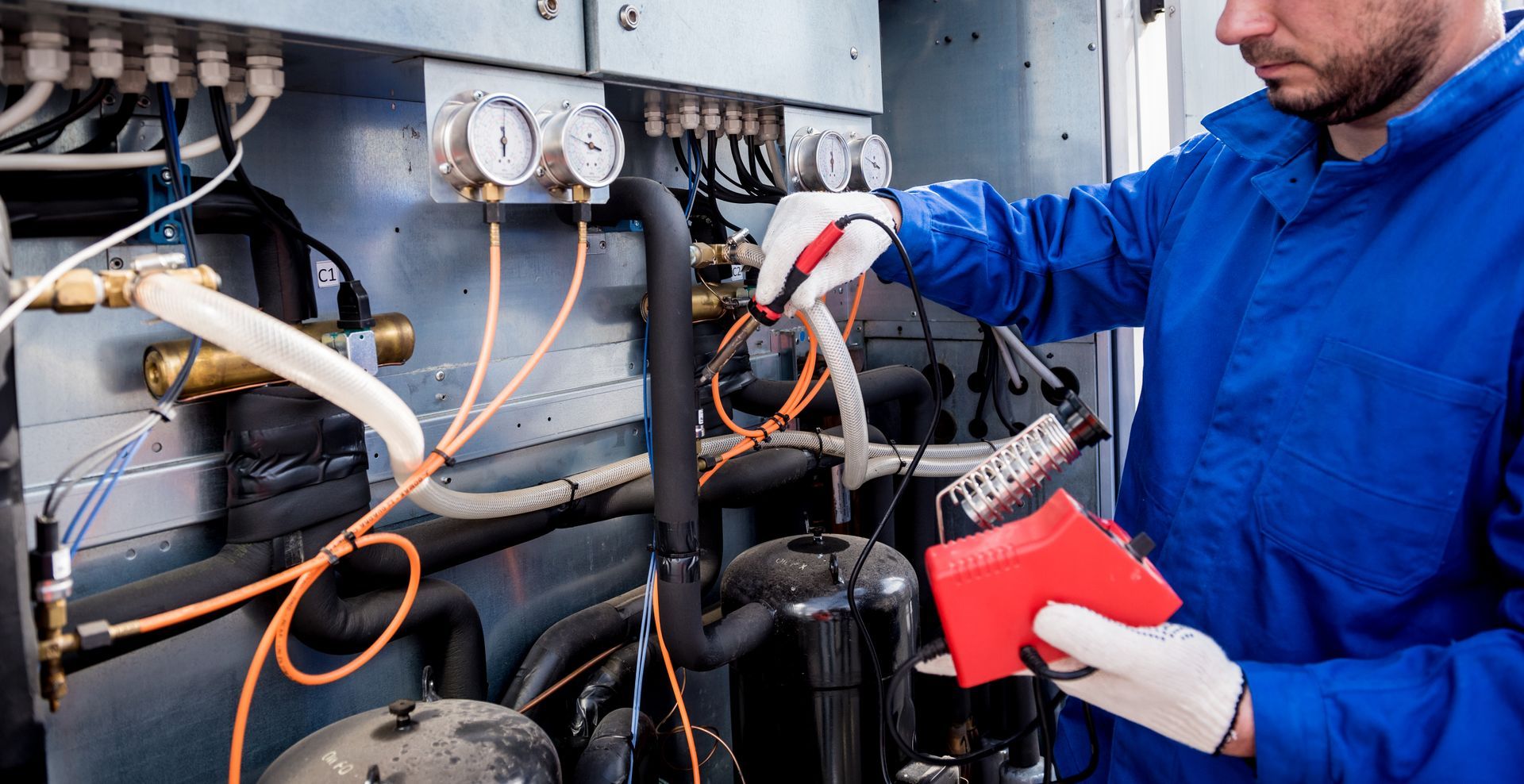 A technician in a blue uniform and gloves uses a red electronic tester to inspect equipment wiring in a utility panel.