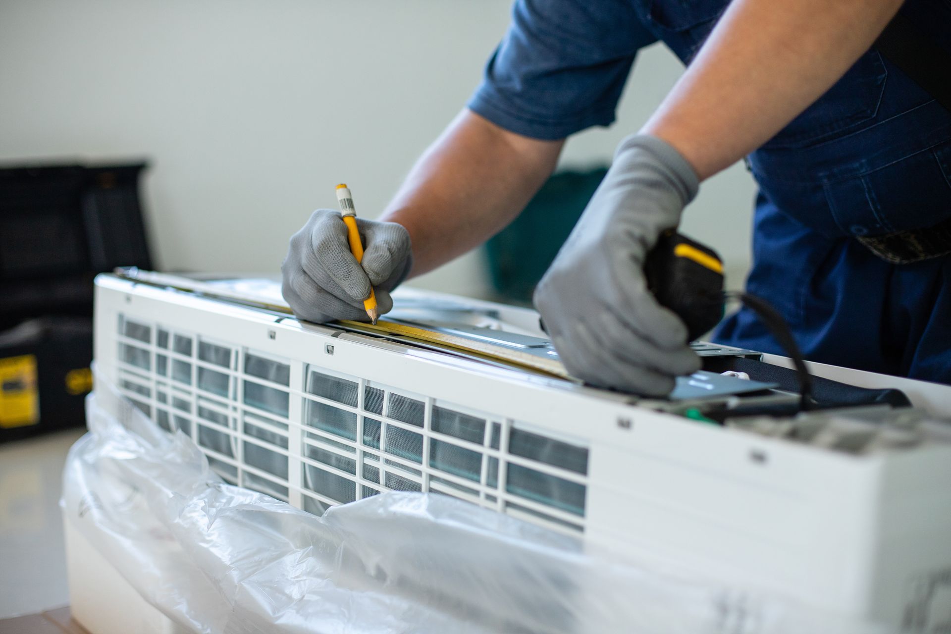 A technician in work gloves measures an air conditioning unit with a tape measure and marks it with a pencil.