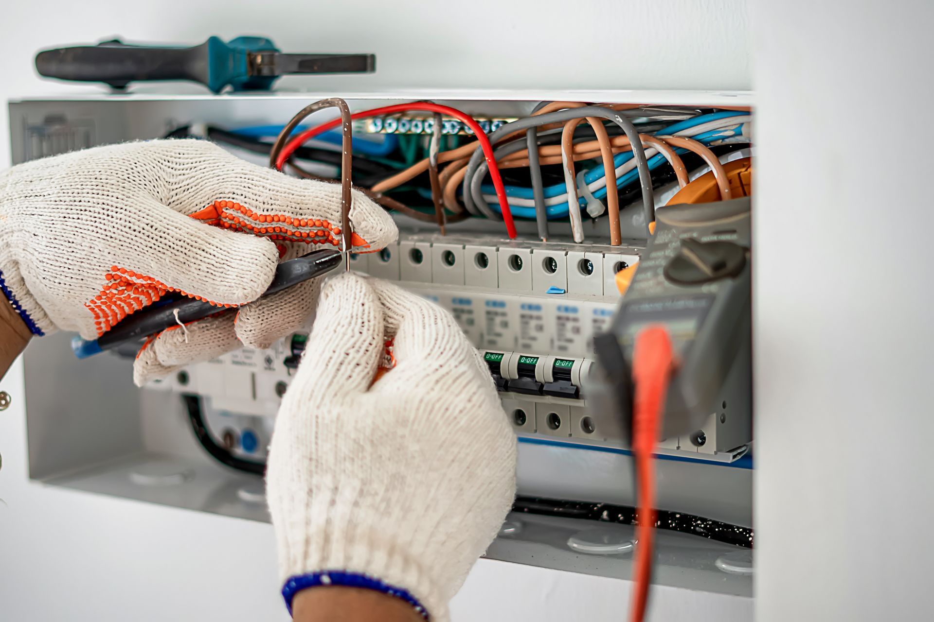 An electrician wearing work gloves uses tools to wire an open electrical breaker panel.