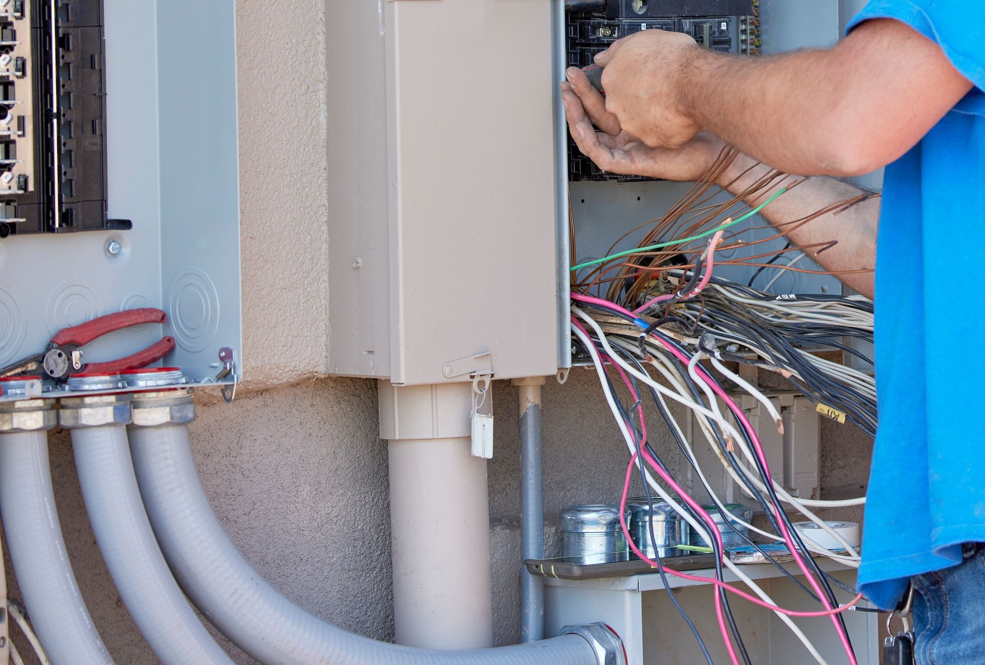 An electrician in a blue shirt works on electrical wiring inside an open outdoor utility panel.